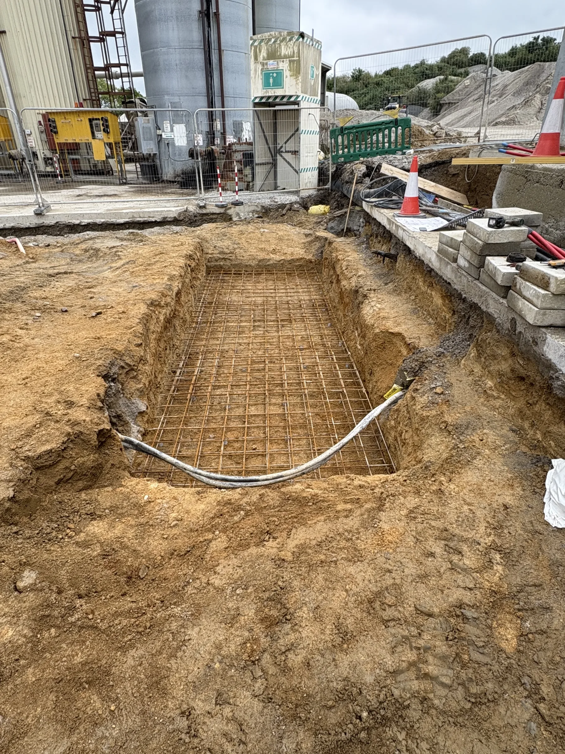 Rectangular excavation containing steel reinforcement mesh and a curved grey conduit, with stacked paving blocks and traffic cones alongside.