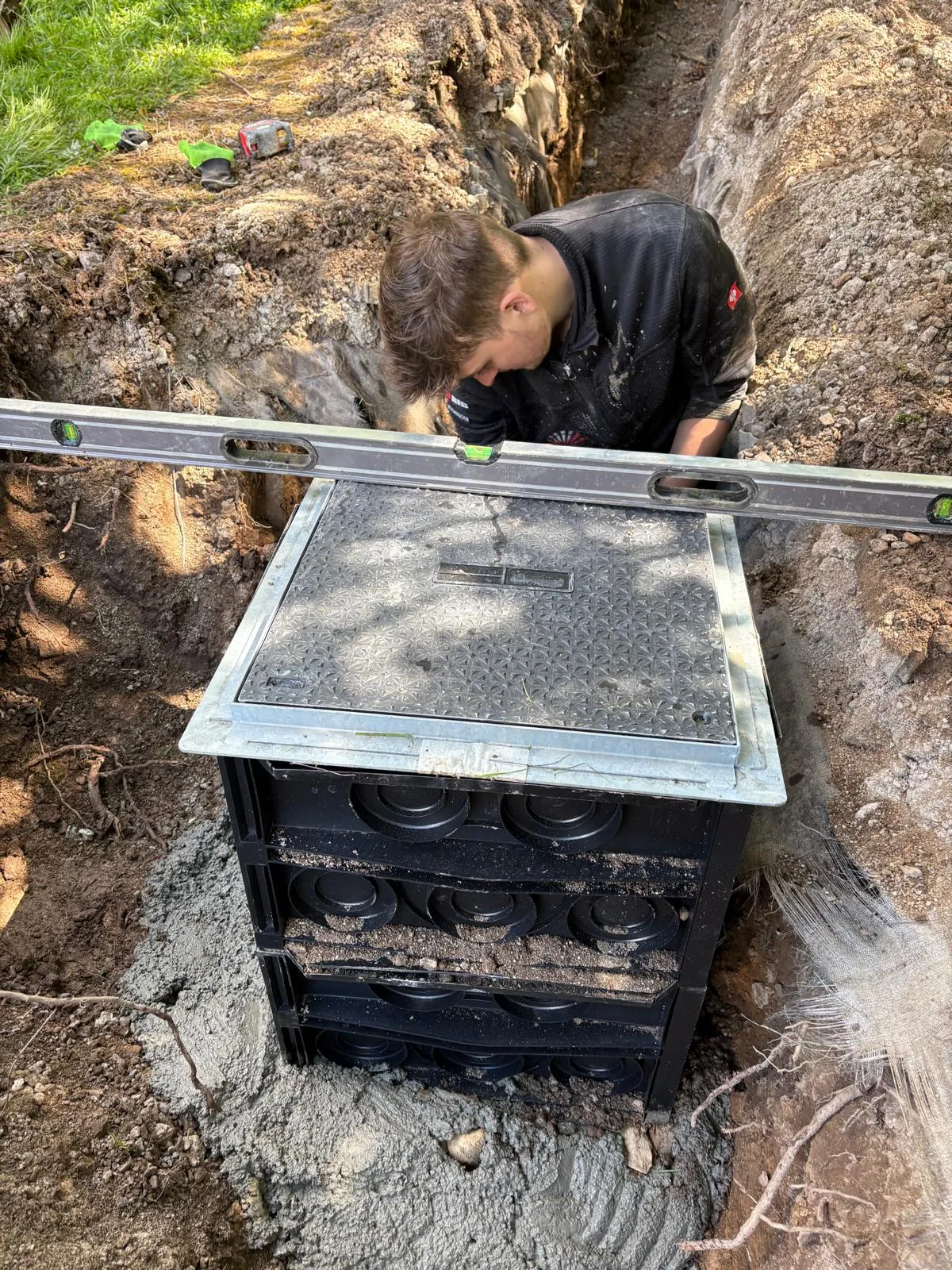 Worker standing in a narrow trench behind a black chamber while a long spirit level rests across its square lid.