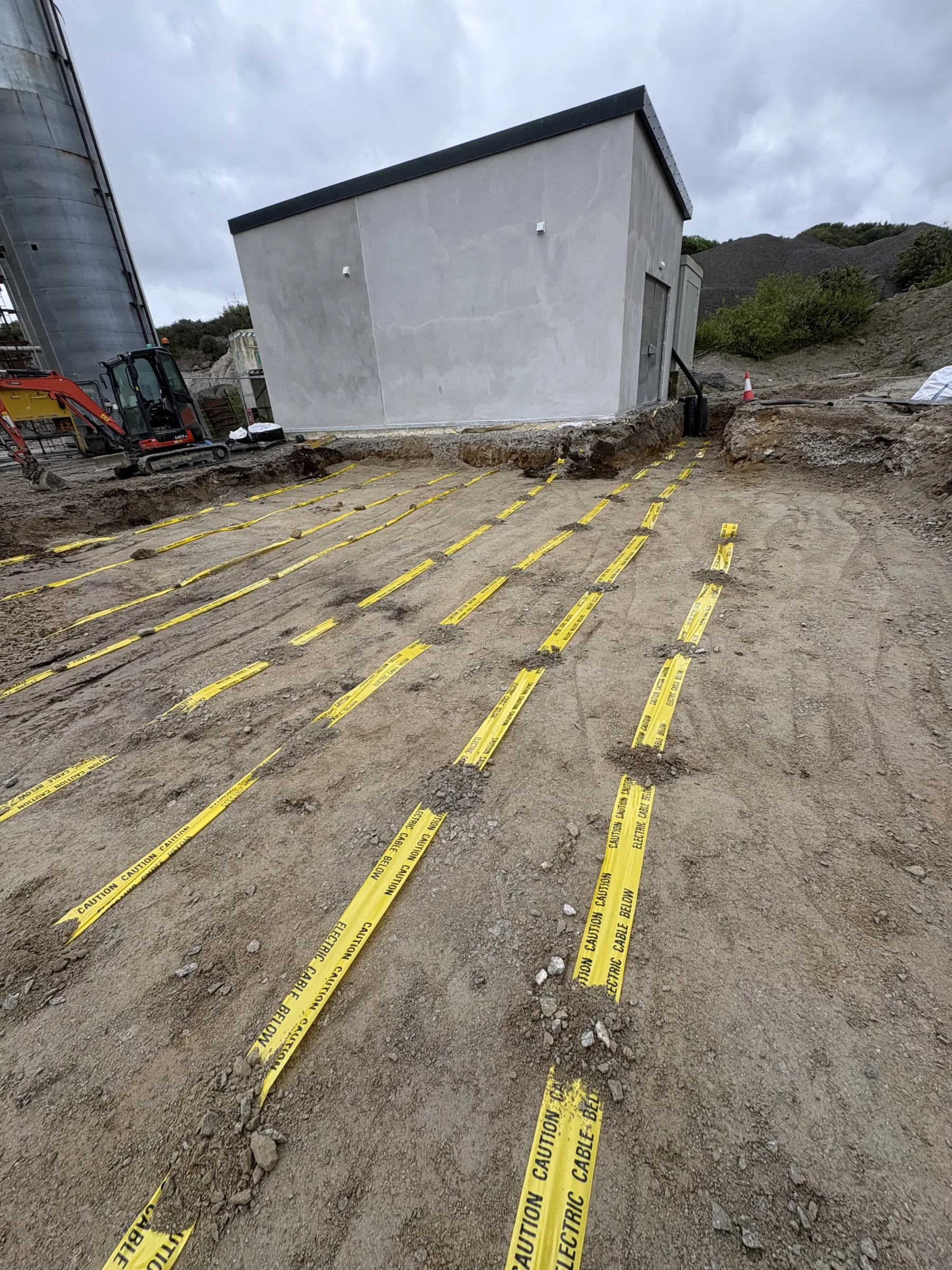 Multiple parallel lines of yellow electrical cable warning tape laid across prepared ground beside a rendered utility building.