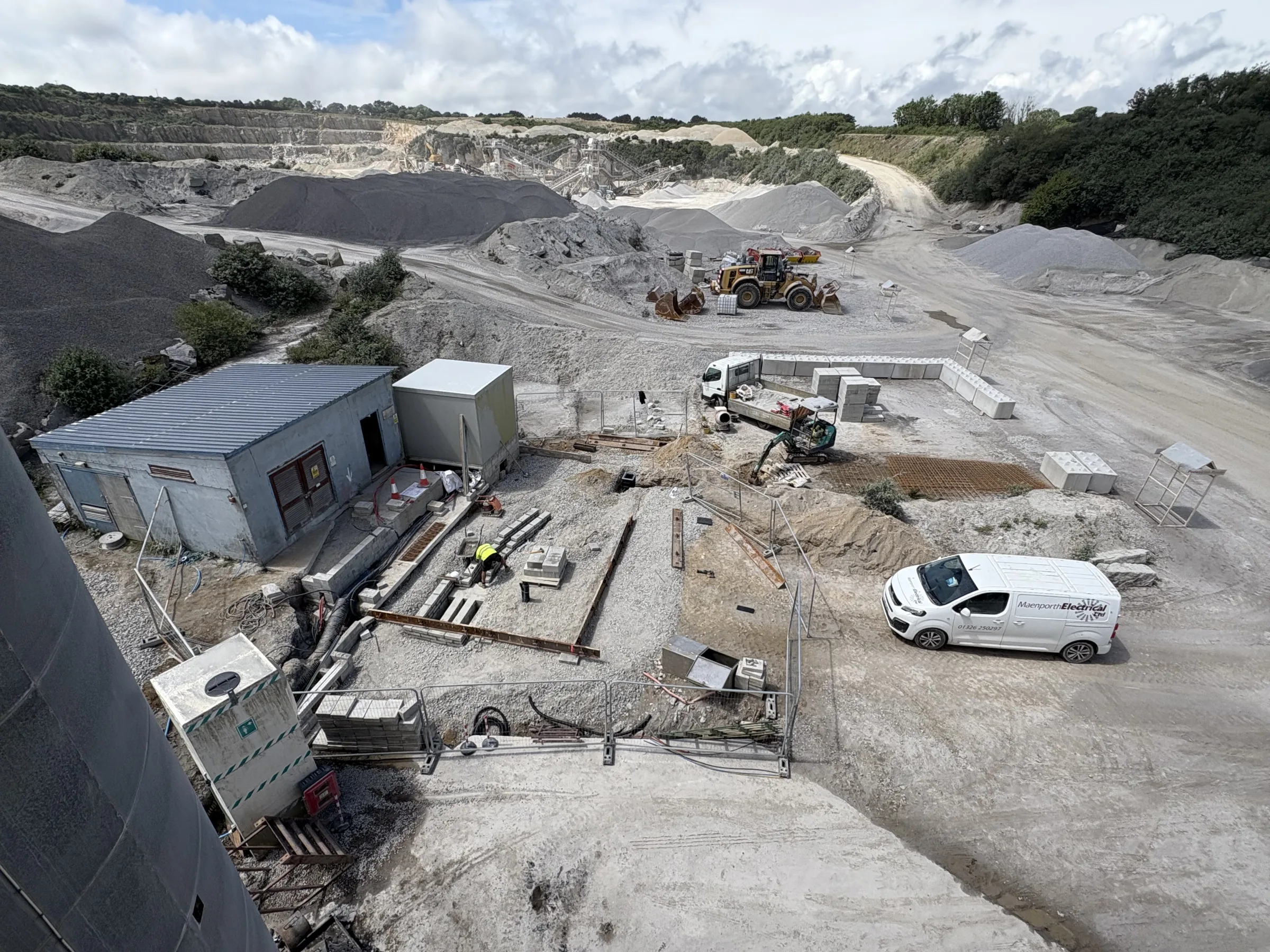 Elevated view of a fenced quarry compound with foundation trenches, workers, plant machinery and two white vans.