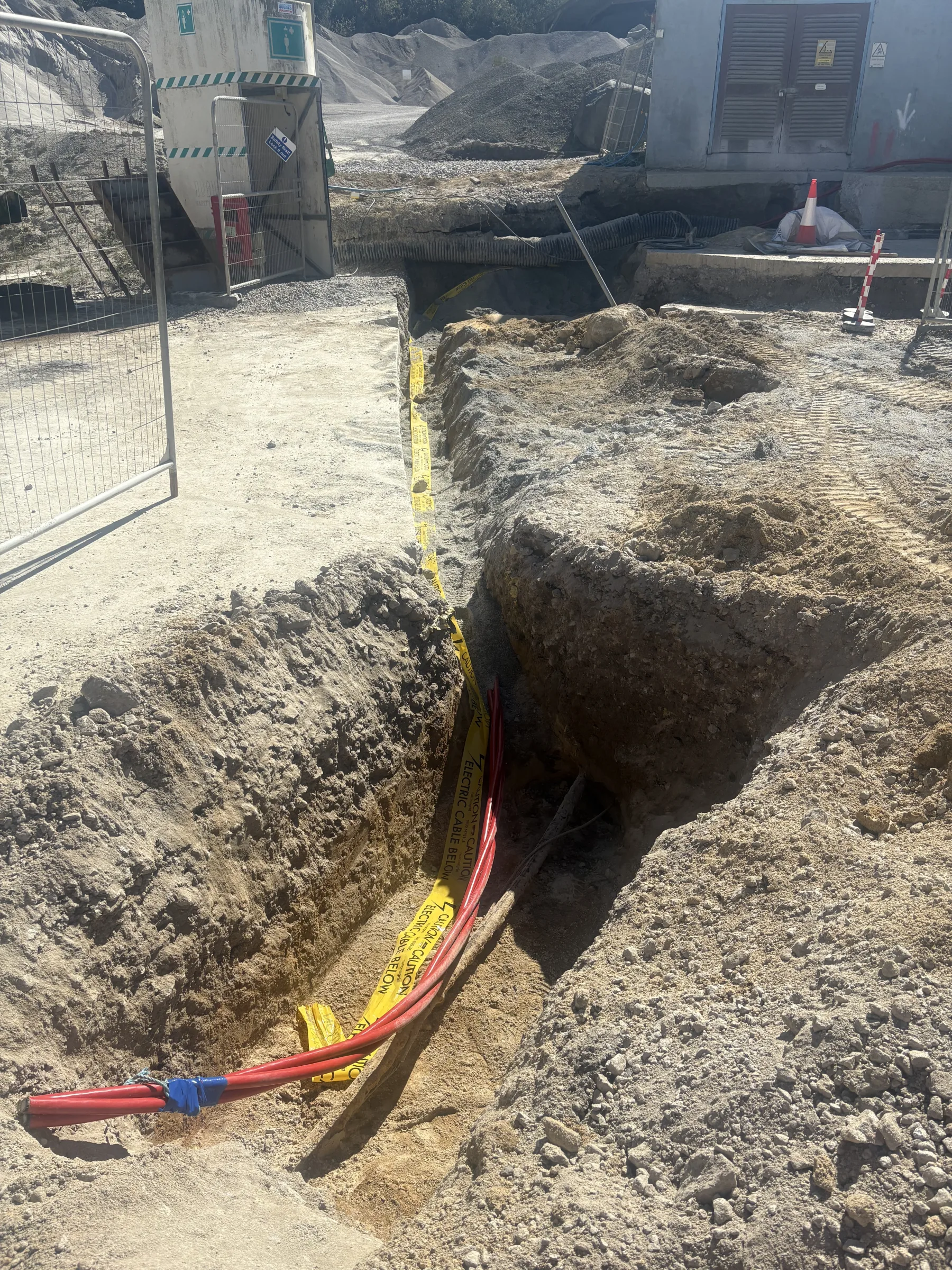 Open trench containing several red ducts, yellow warning tape and a timber board, surrounded by excavated sandy ground.