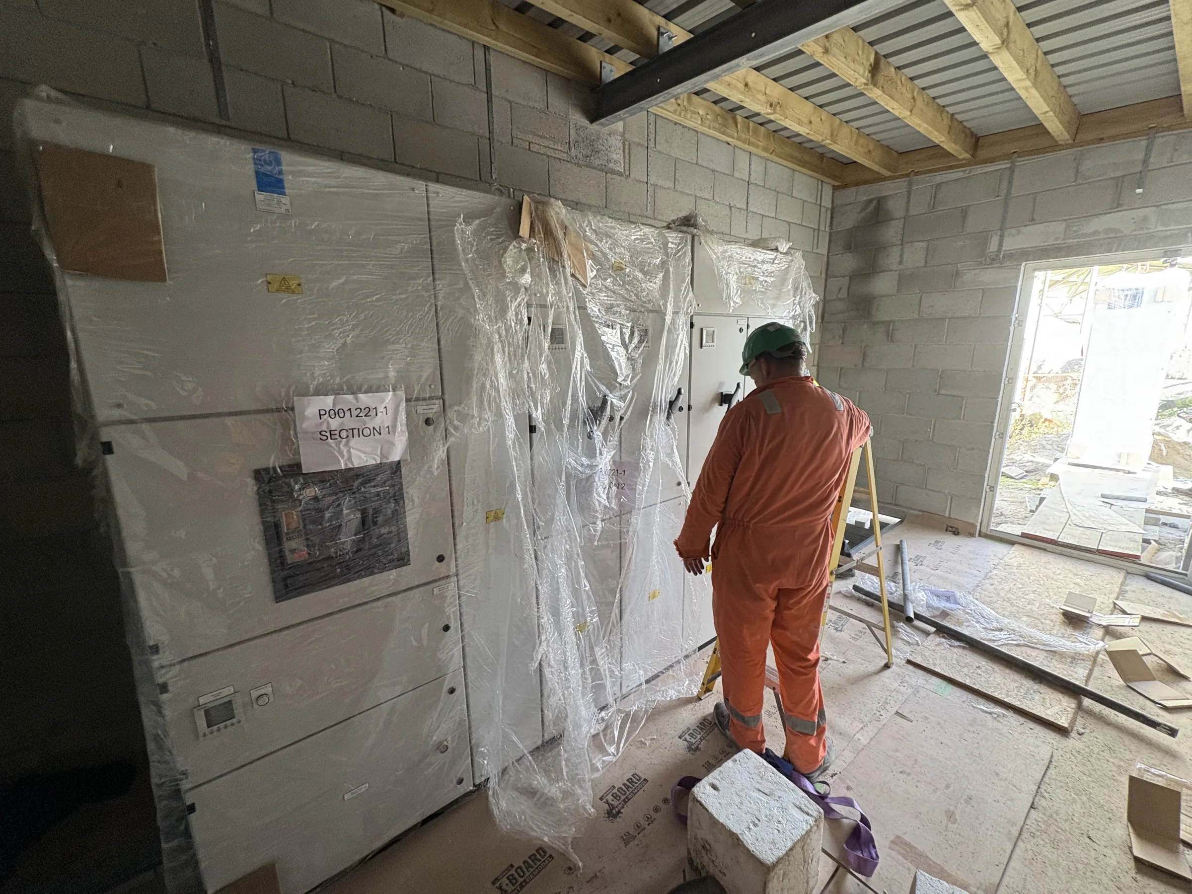 Worker in orange protective clothing and a green hard hat standing beside large wrapped electrical equipment inside an unfinished blockwork building.