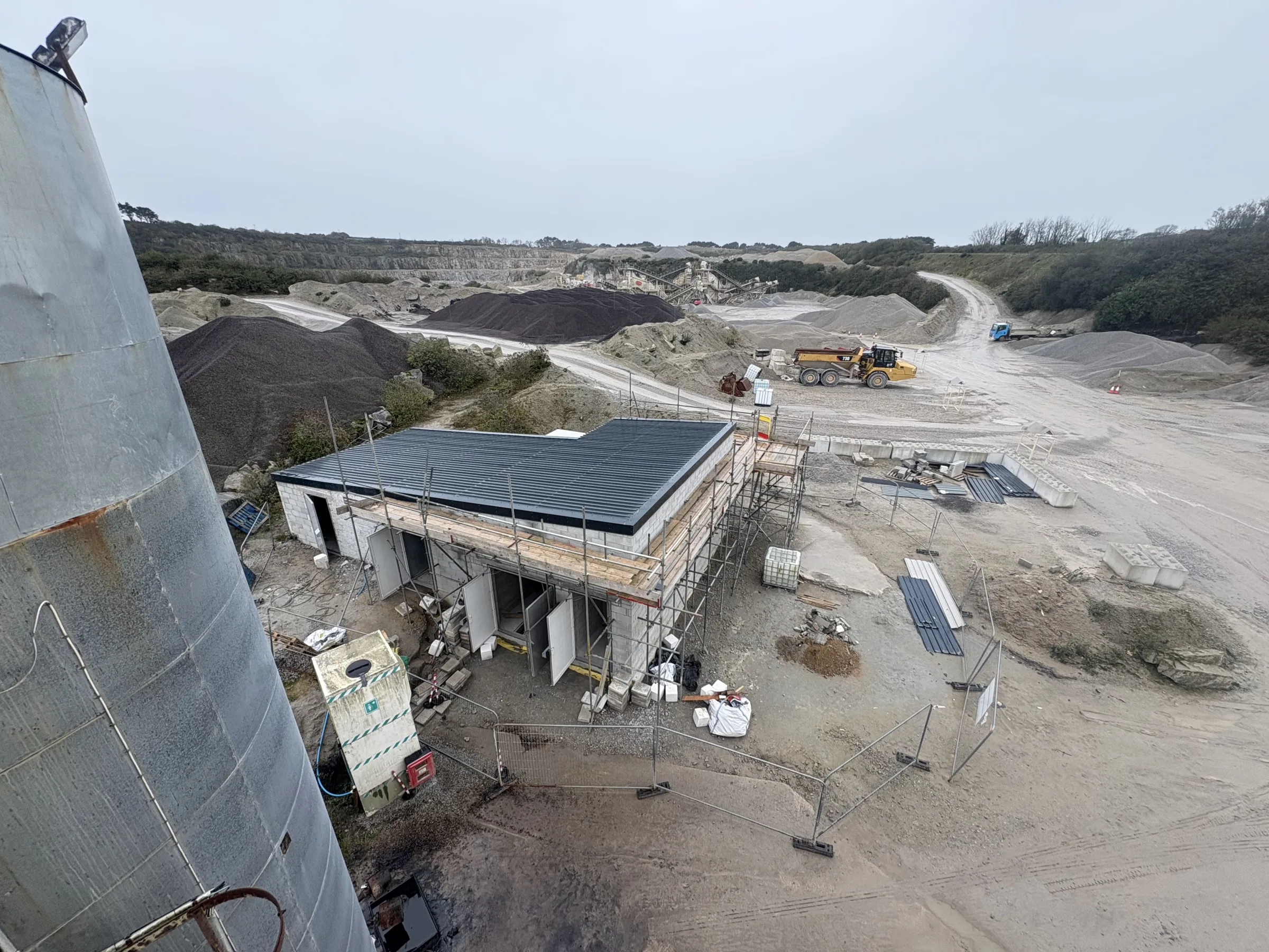Elevated view across a quarry-like construction site with a small scaffolded building, striped roof covering, aggregate piles and machinery.