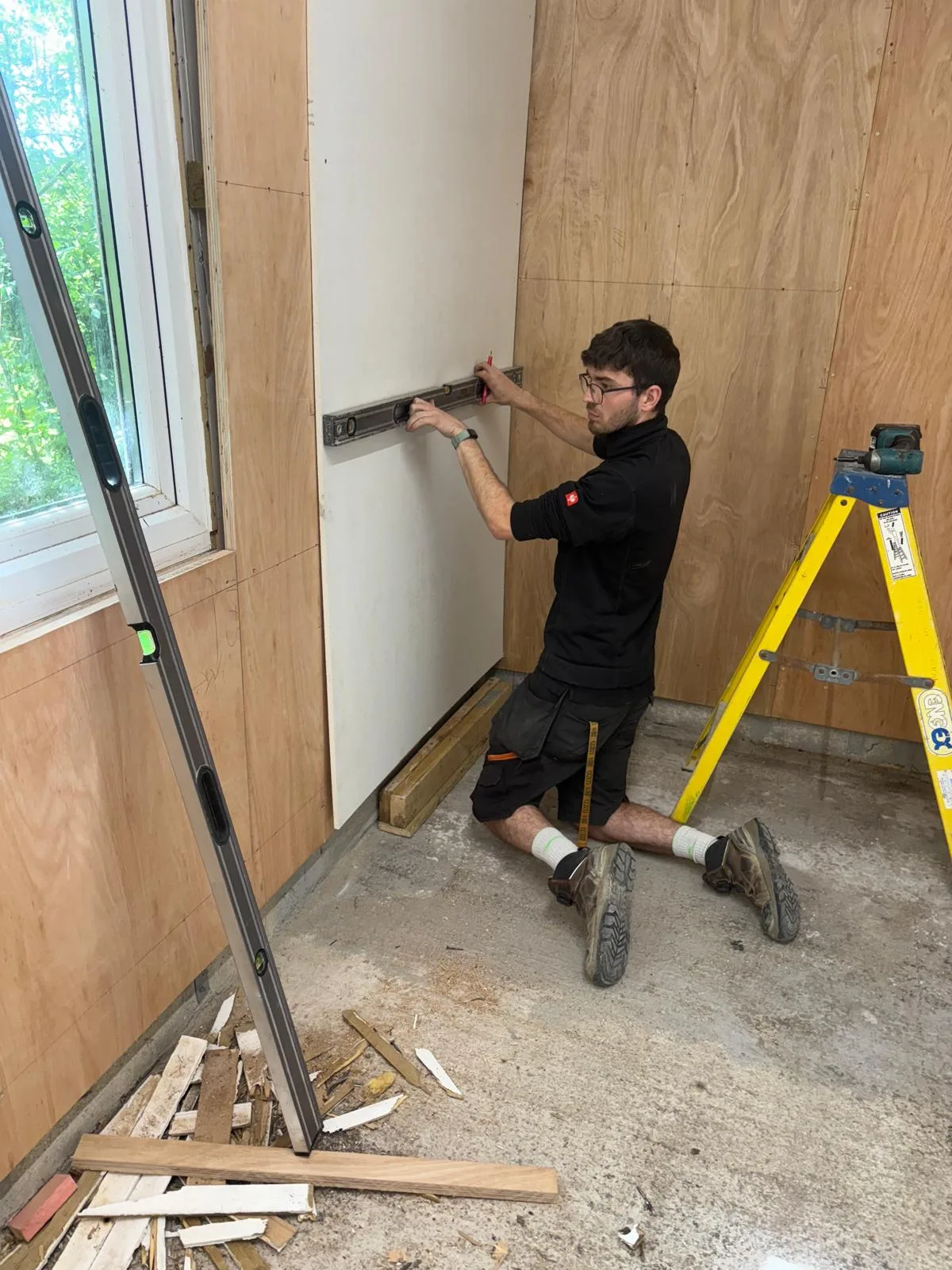 Electrician kneeling beside a wall and fitting a long metal electrical back box, with plywood wall panels, a window, spirit level and yellow stepladder visible.