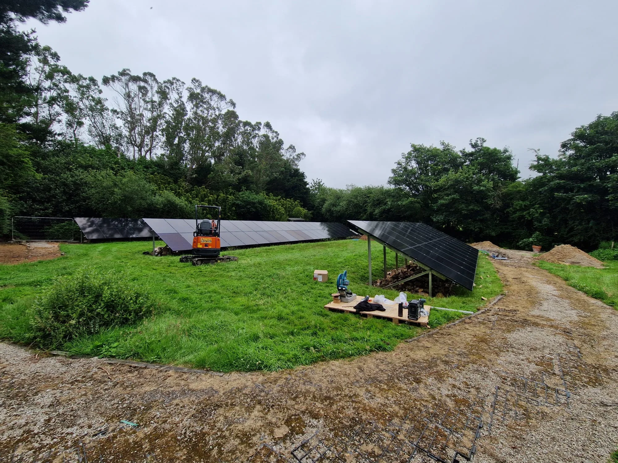 Rows of ground-mounted solar panels across a grassy clearing, with a small orange excavator, tools and a gravel track.