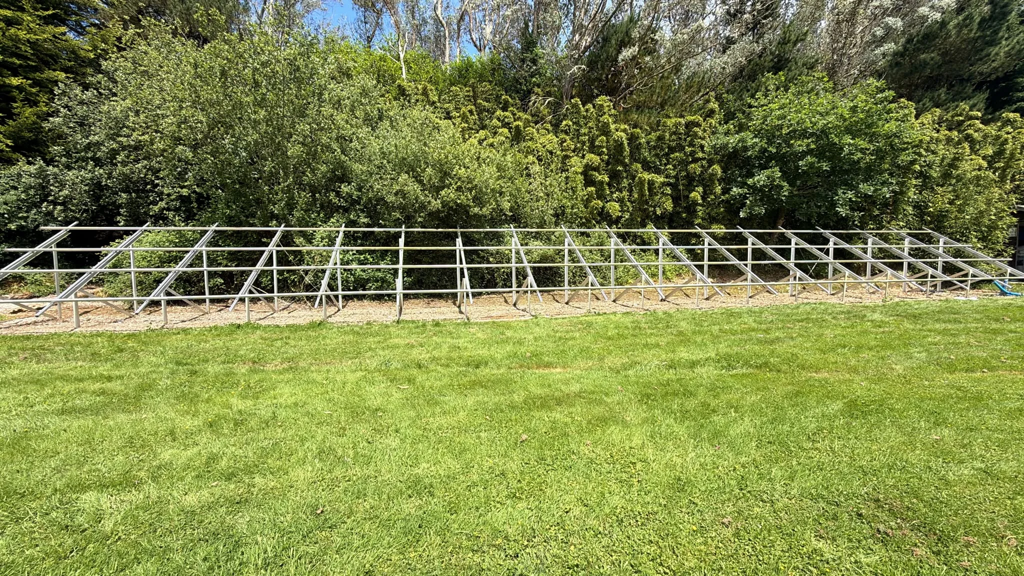 Long metal mounting framework without panels installed, positioned on grass in front of dense trees and shrubs.