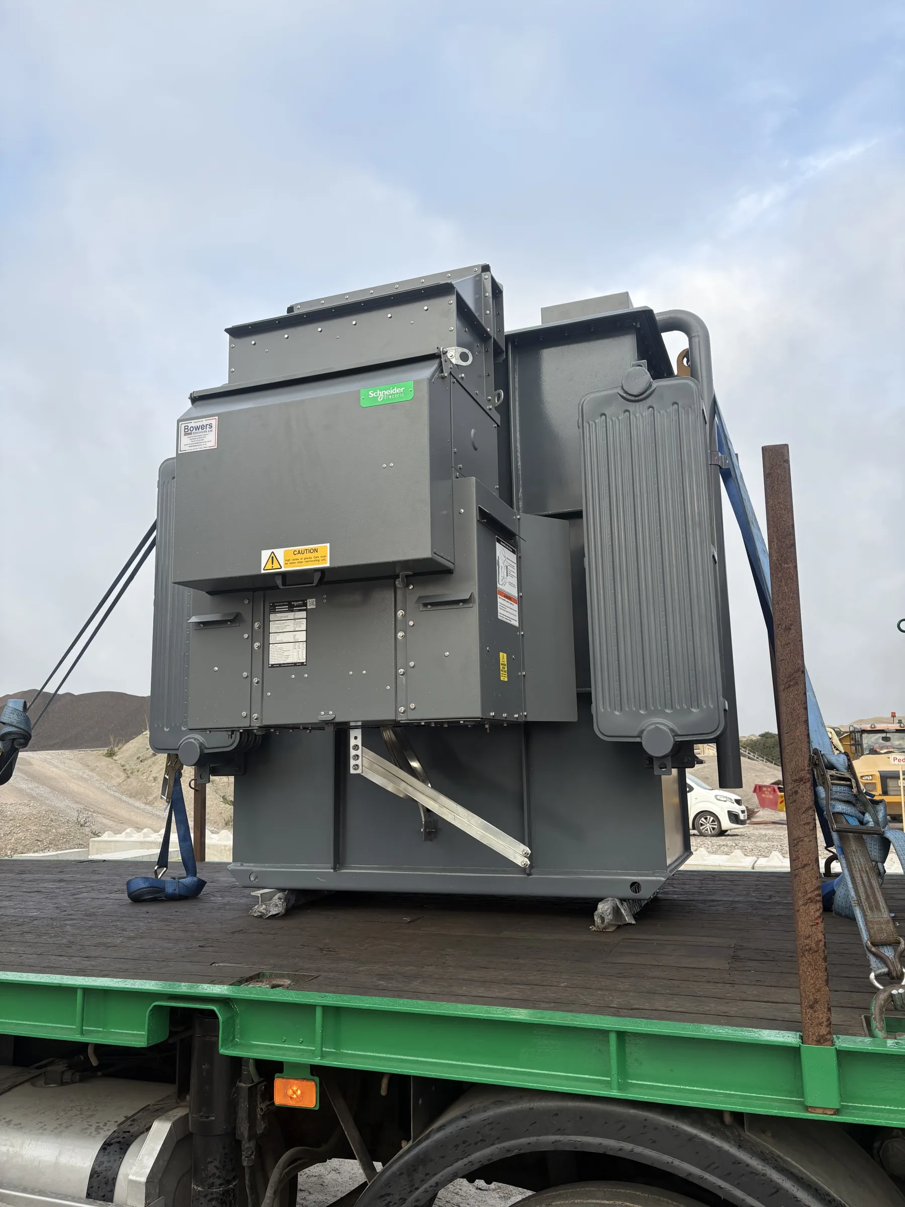 Large grey transformer secured with blue straps on the bed of a green flatbed lorry at an outdoor work site.