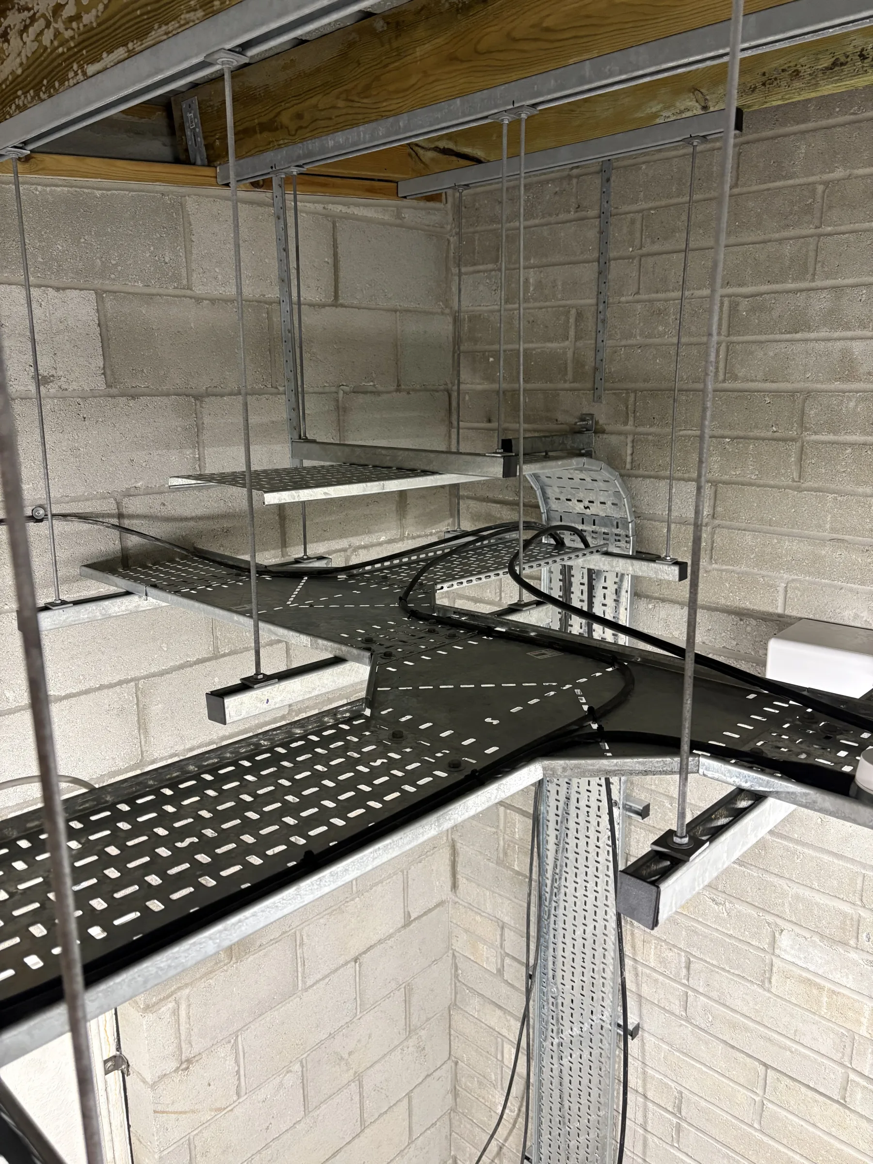 Black perforated cable trays and silver metal containment suspended from the ceiling beside unfinished block walls.