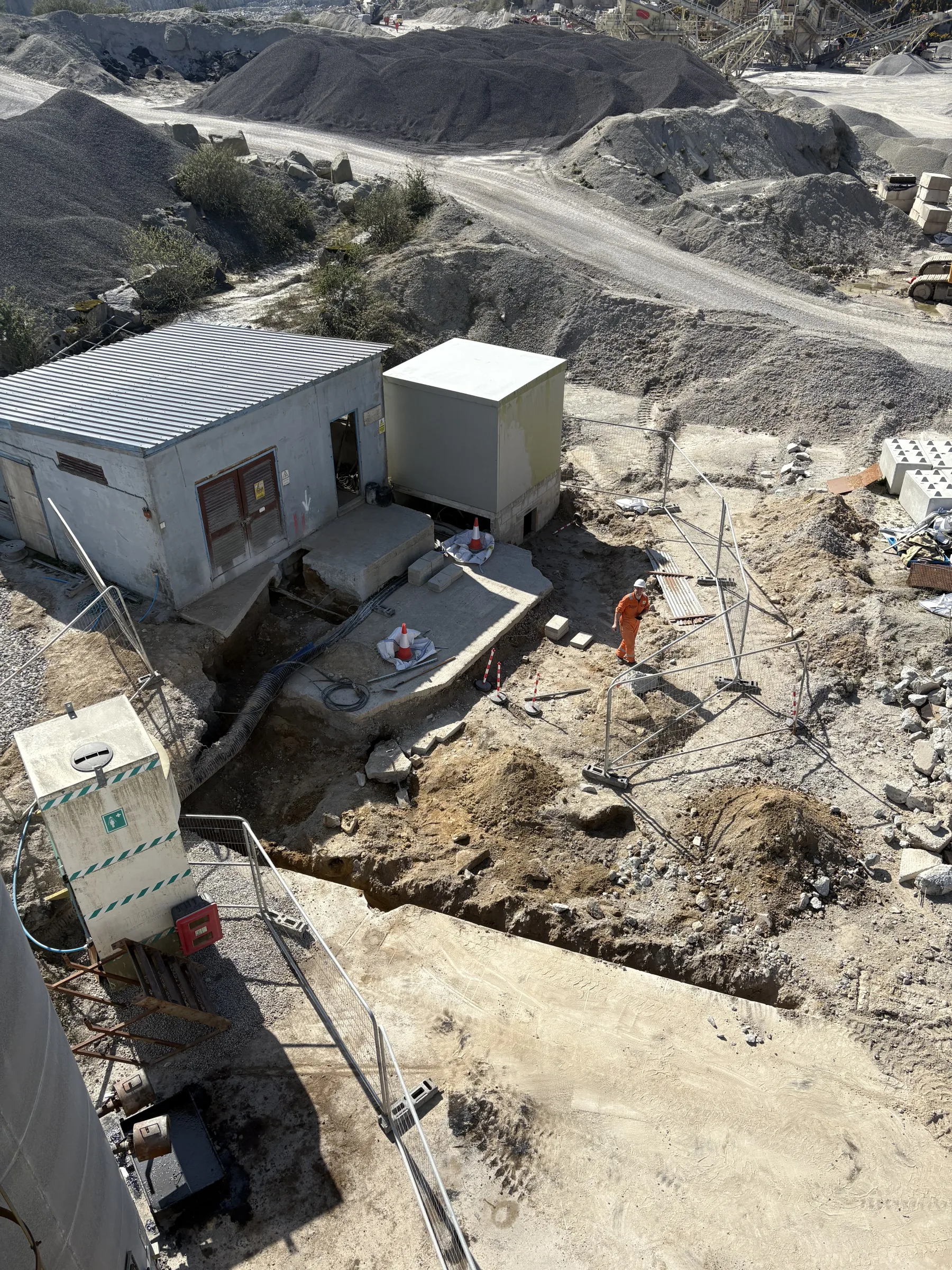 Elevated view over an industrial site with a small building, a large grey enclosure, open excavations, barriers and aggregate stockpiles.