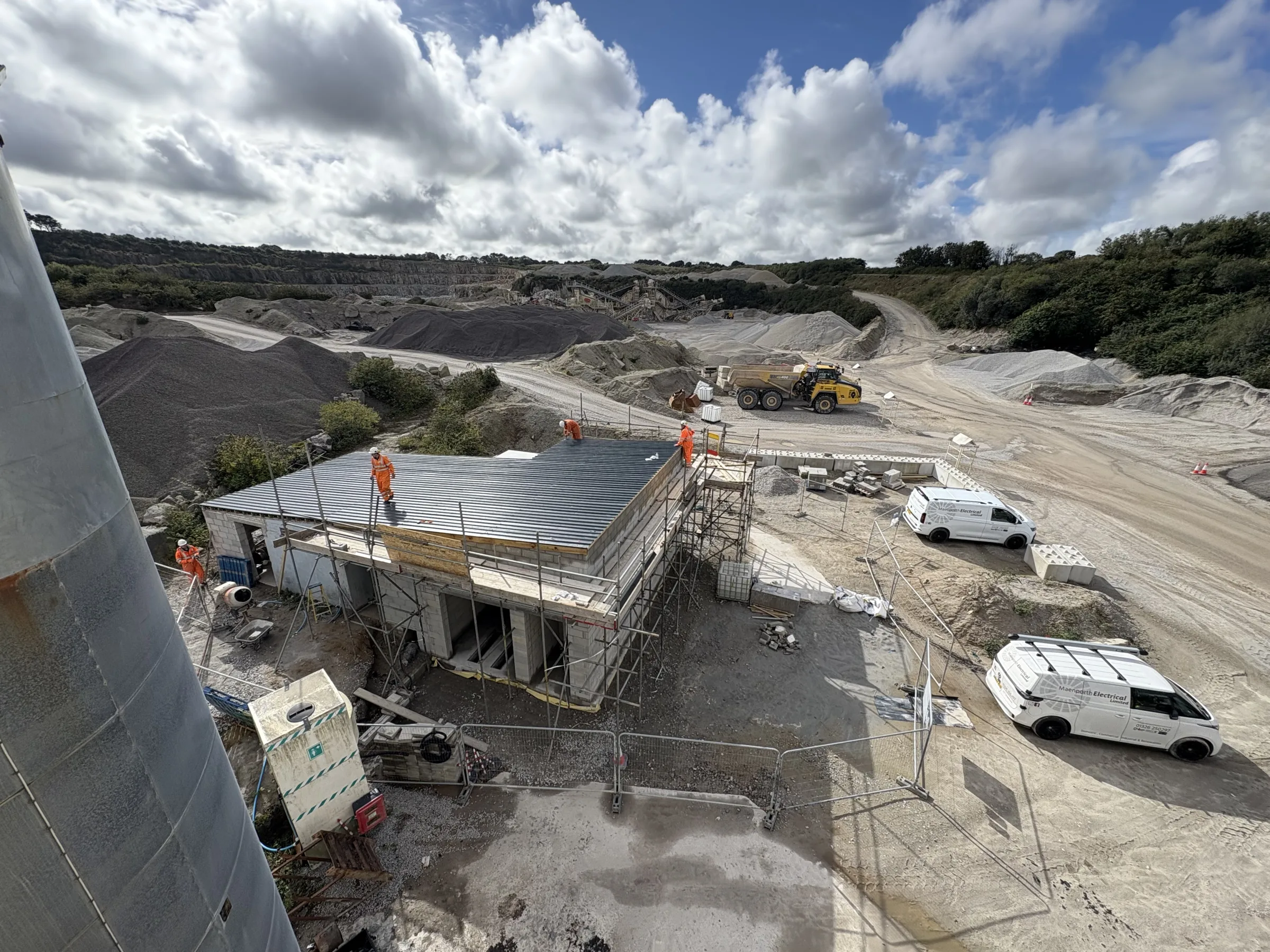 Elevated view of workers in orange clothing on the striped roof of a scaffolded building, with two white vans and a yellow dump truck nearby.