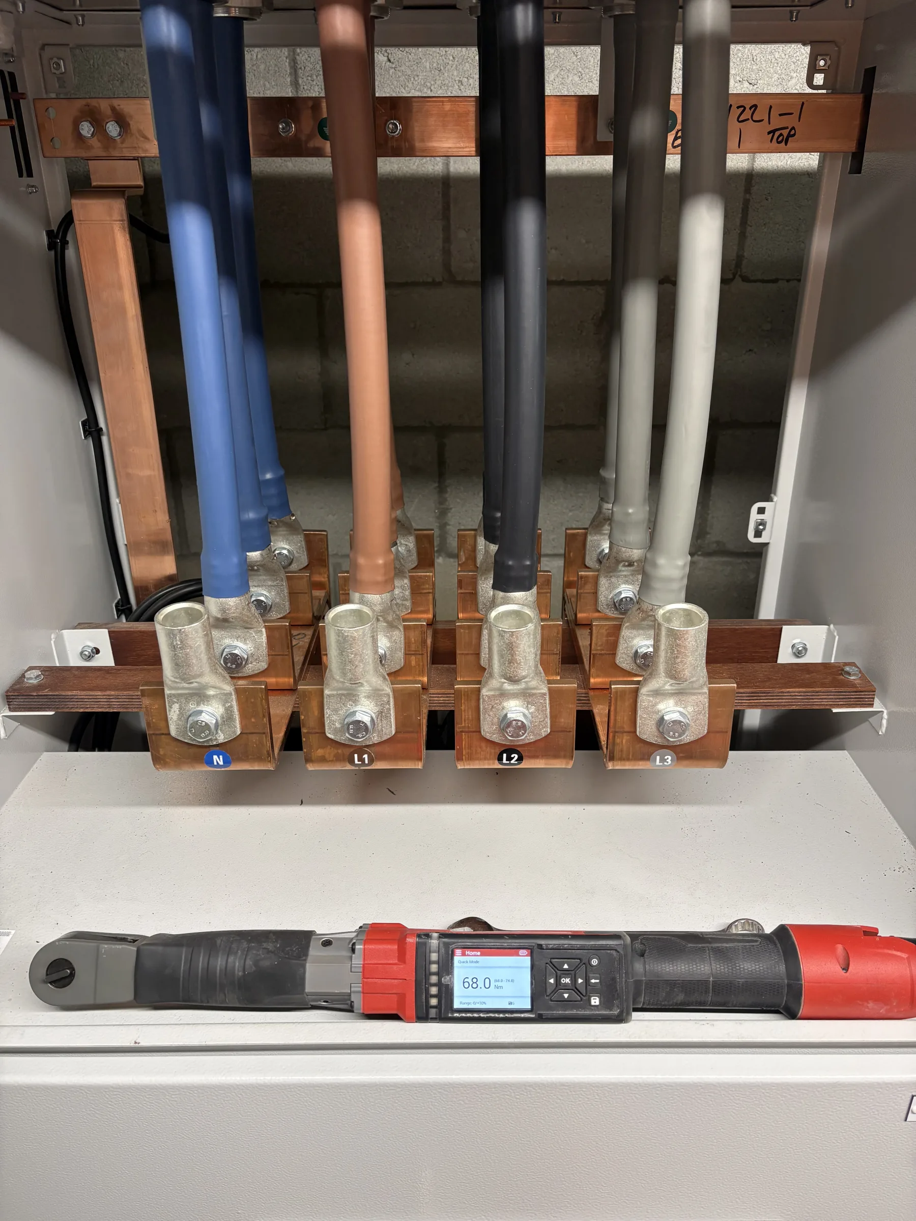 Open electrical cabinet containing five large coloured cables connected to copper bars, with a digital hand tool resting below.