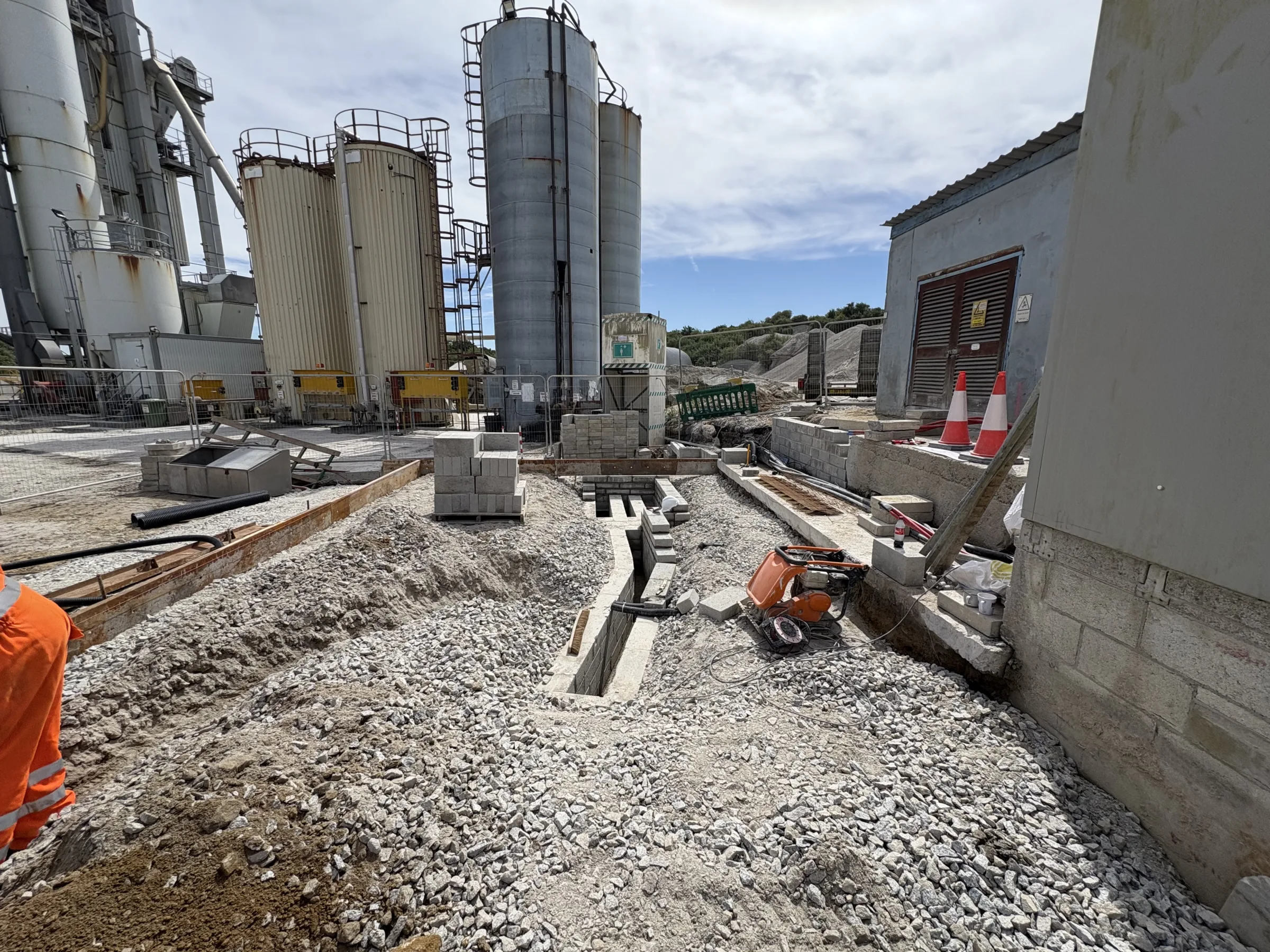 Gravel-covered construction area between industrial buildings and silos, with shallow trenches, concrete blocks and a small compactor.