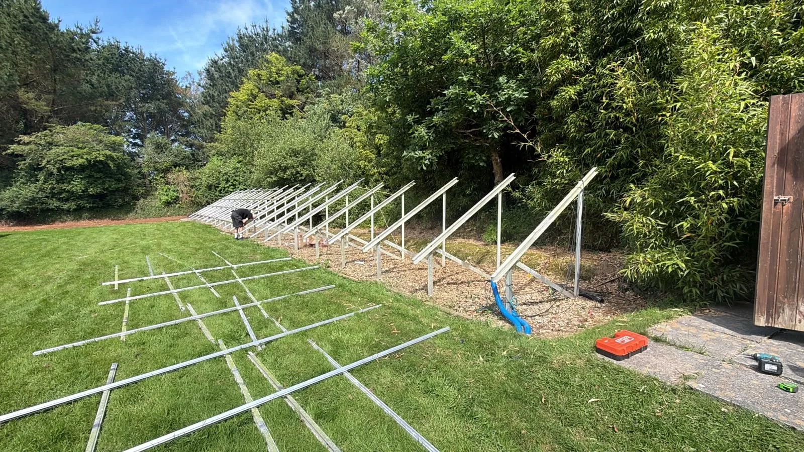 Angled metal solar mounting posts installed along a gravel strip beside trees, with unassembled frame sections laid out on the grass and tools nearby.