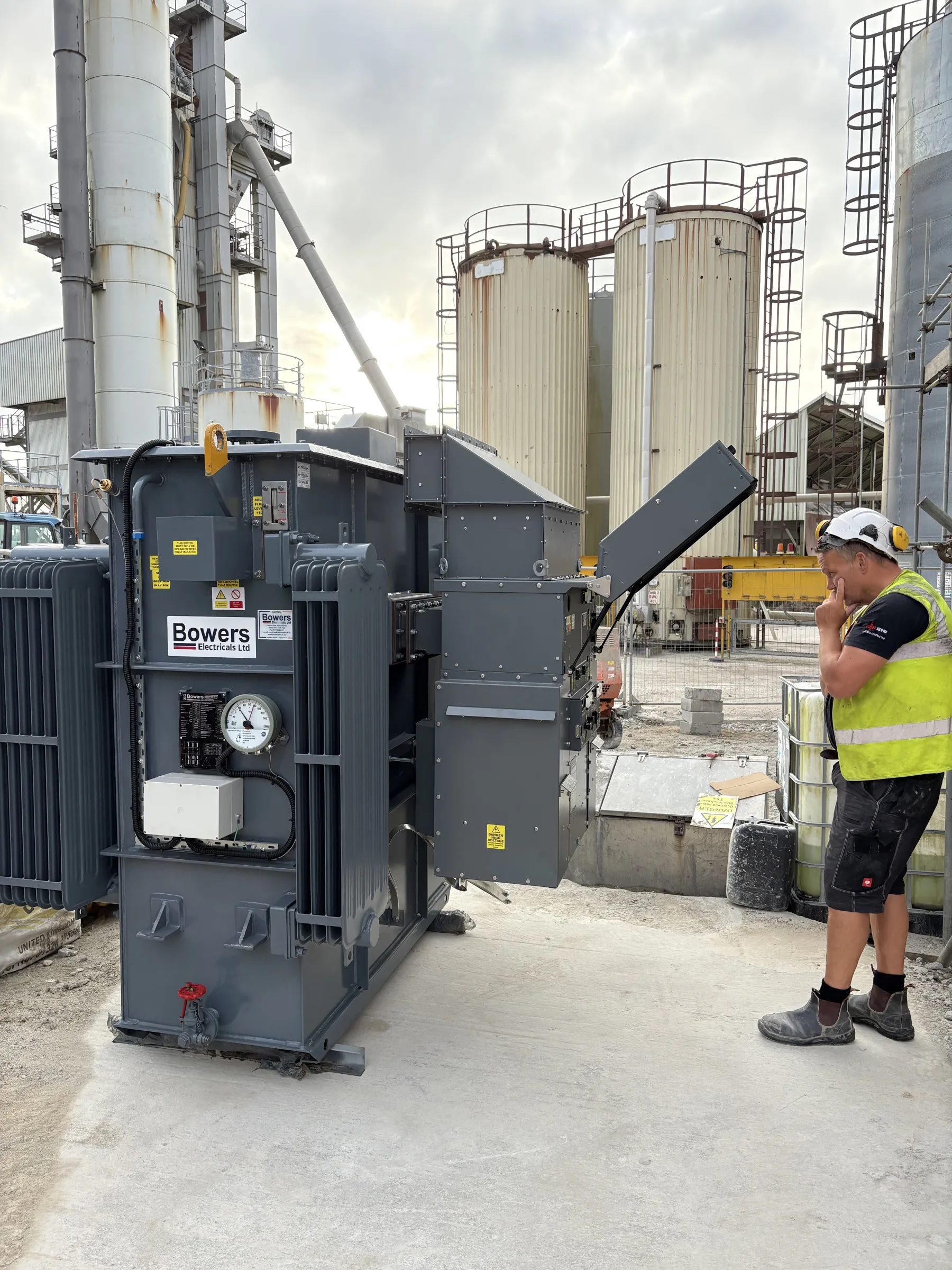 Large grey transformer standing beside a concrete base at an industrial site, with a worker wearing a hard hat and high-visibility vest nearby.