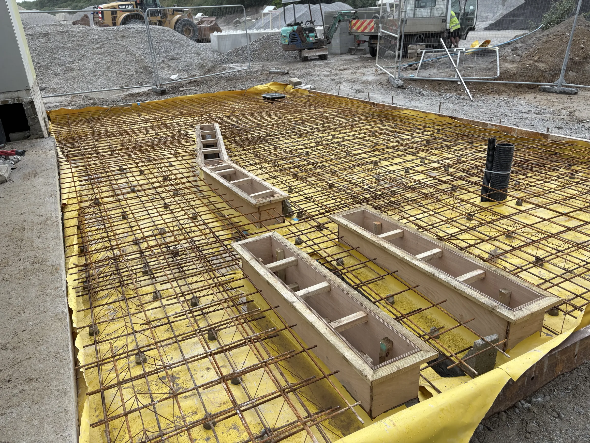 Close view of steel reinforcement mesh over yellow sheeting, with timber service formwork and a black pipe opening.