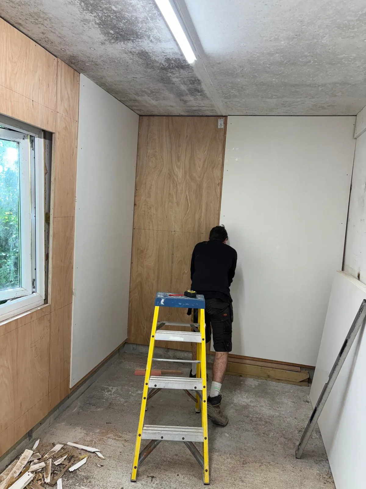 Worker facing a plywood-lined wall in a small room, with a yellow step ladder, window and stacked wall boards visible.