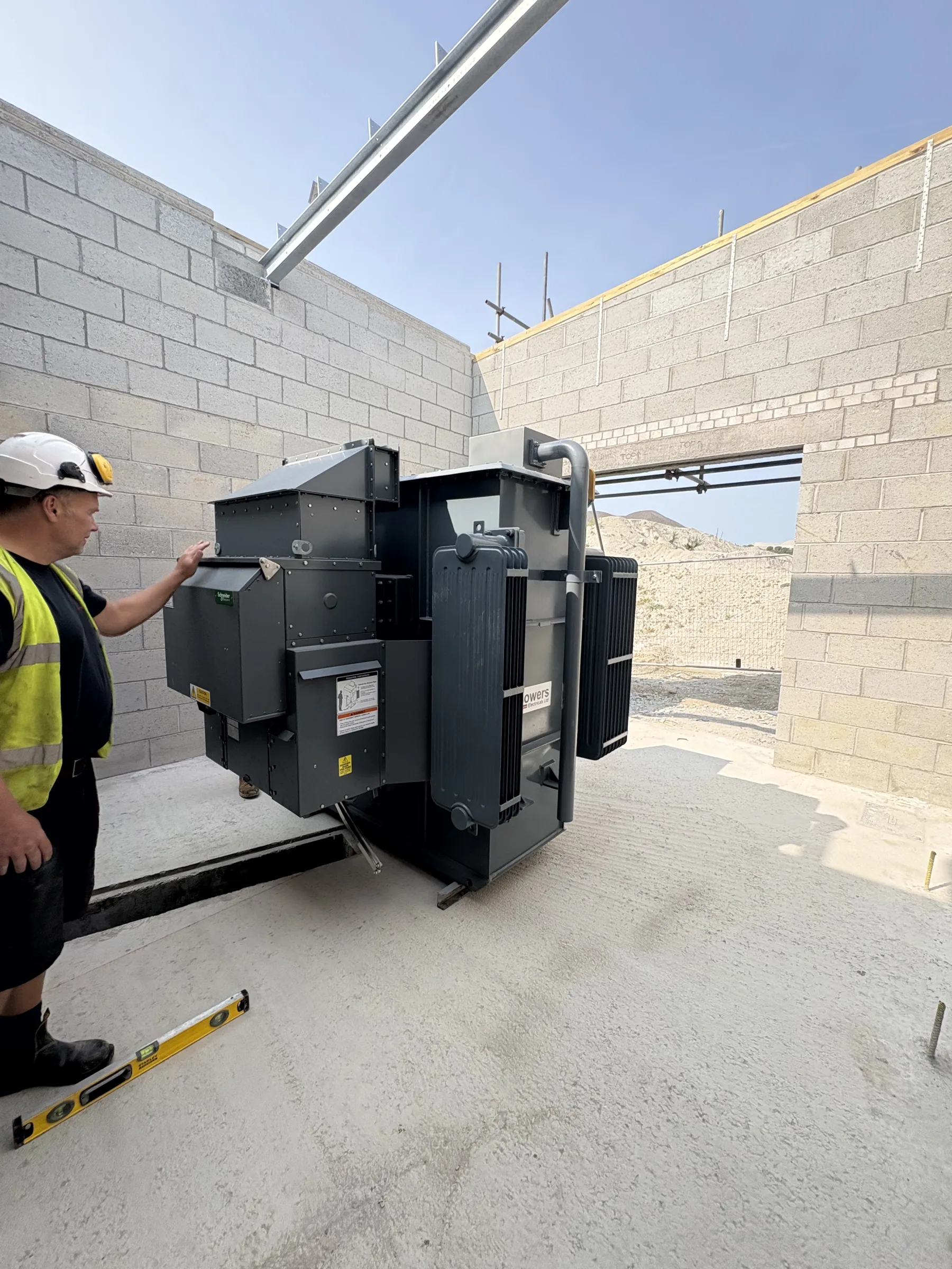 Worker in a hard hat and high-visibility vest standing beside a large black electrical transformer inside an unfinished blockwork room.