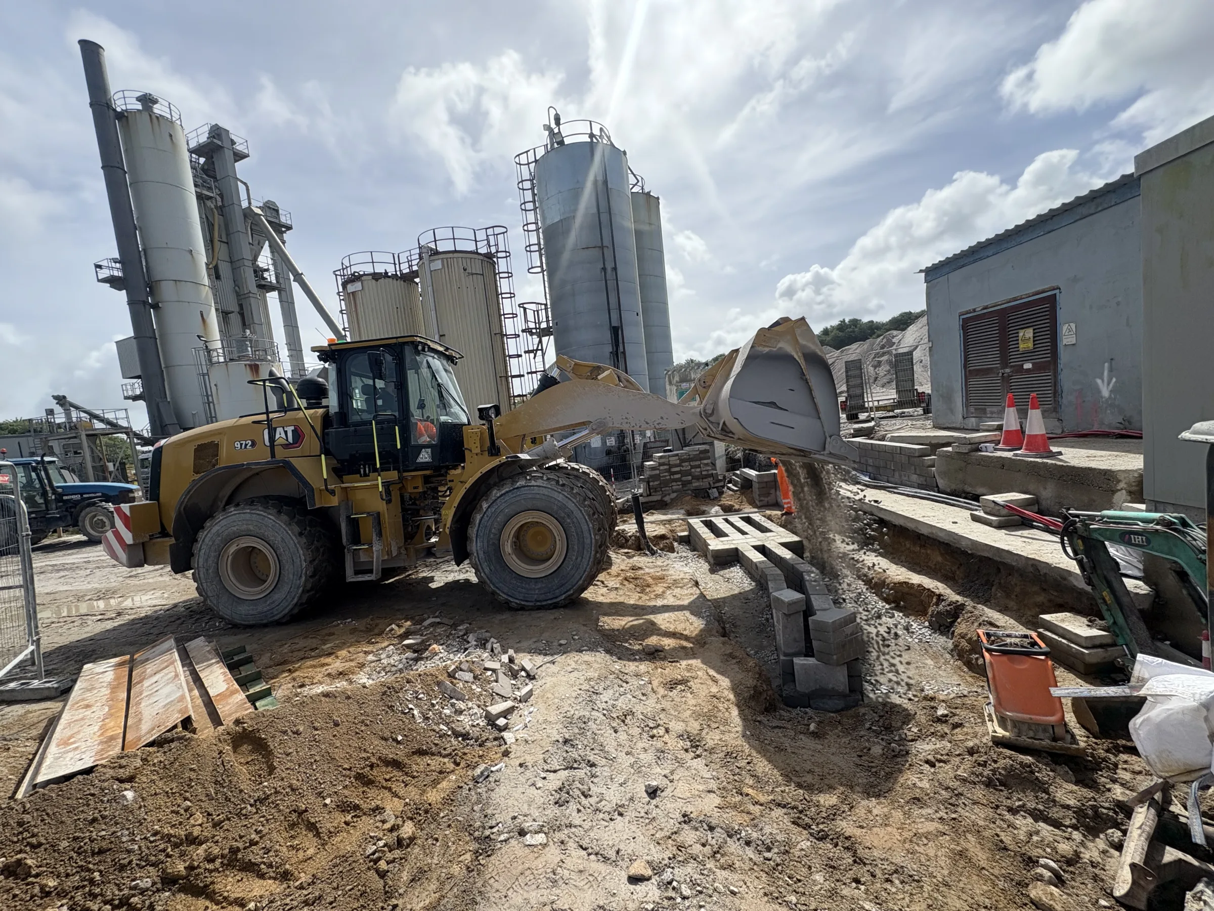 Large wheeled loader with raised bucket beside excavated ground, stacked blocks, barriers and industrial silos.