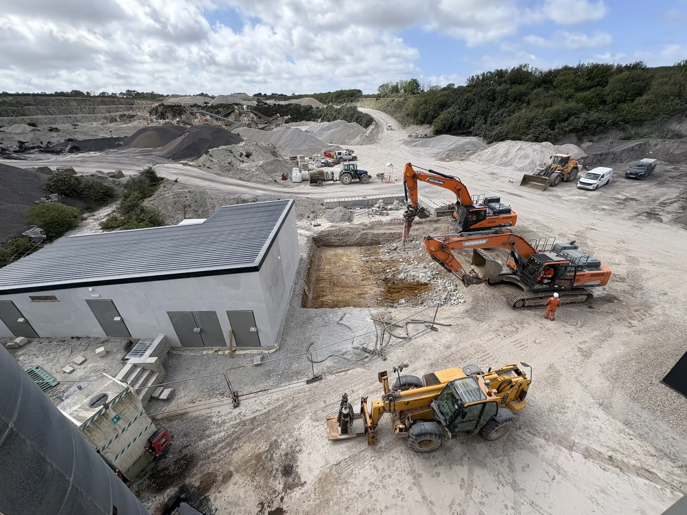 Elevated view of orange excavators and a yellow loader around a rectangular excavation beside a grey industrial building, with aggregate piles in the distance.