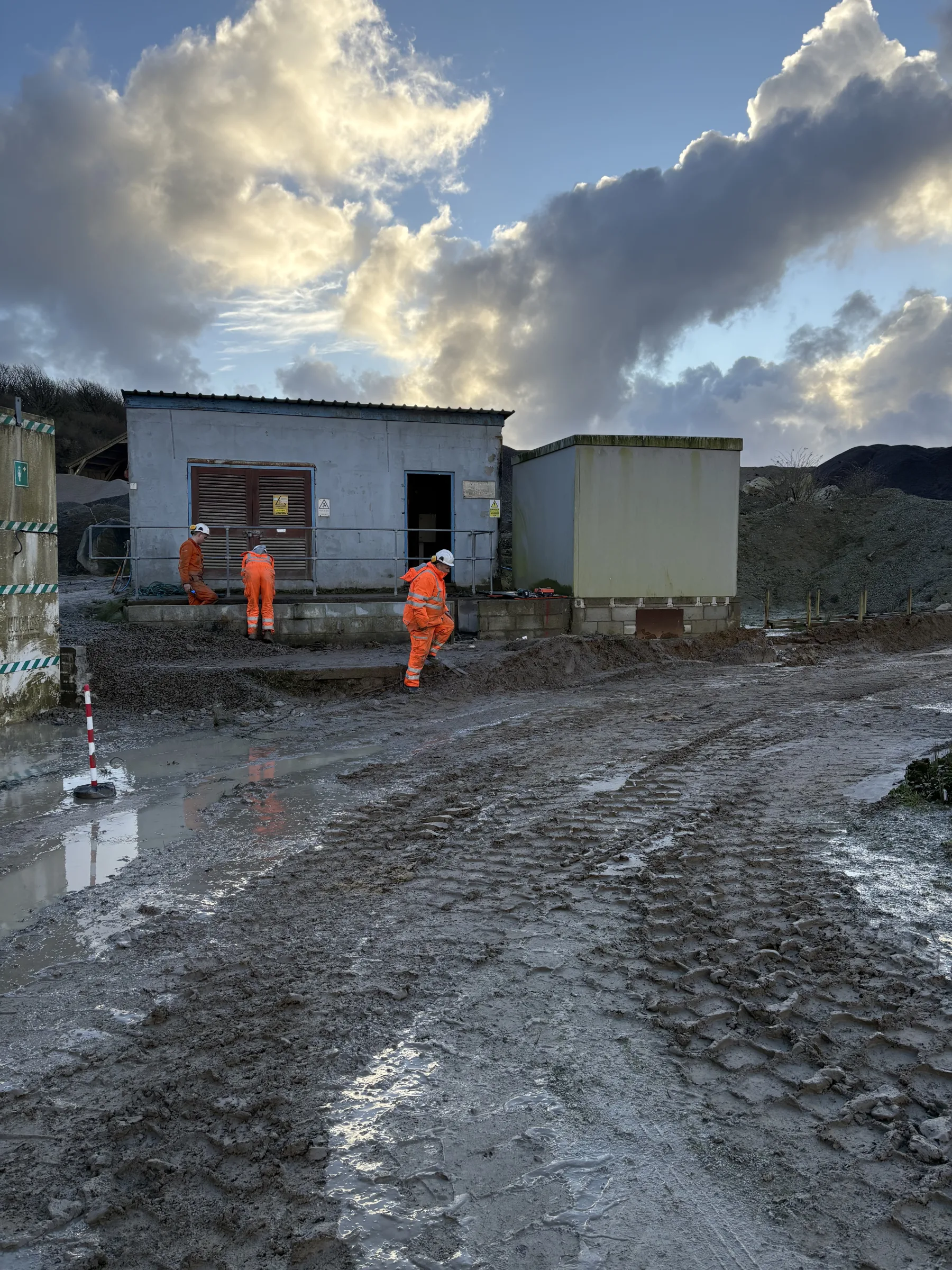 Three workers in orange high-visibility clothing stand beside two utility buildings on a muddy industrial site with puddles and aggregate piles.