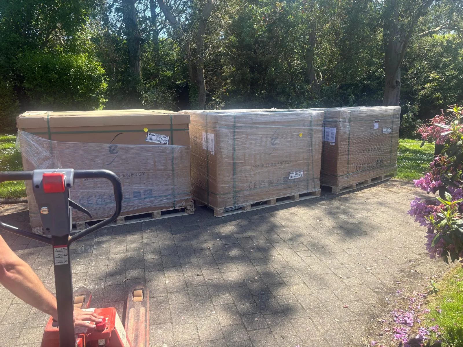 Four large cardboard-wrapped loads on wooden pallets standing on paving, with a red pallet truck in the foreground.