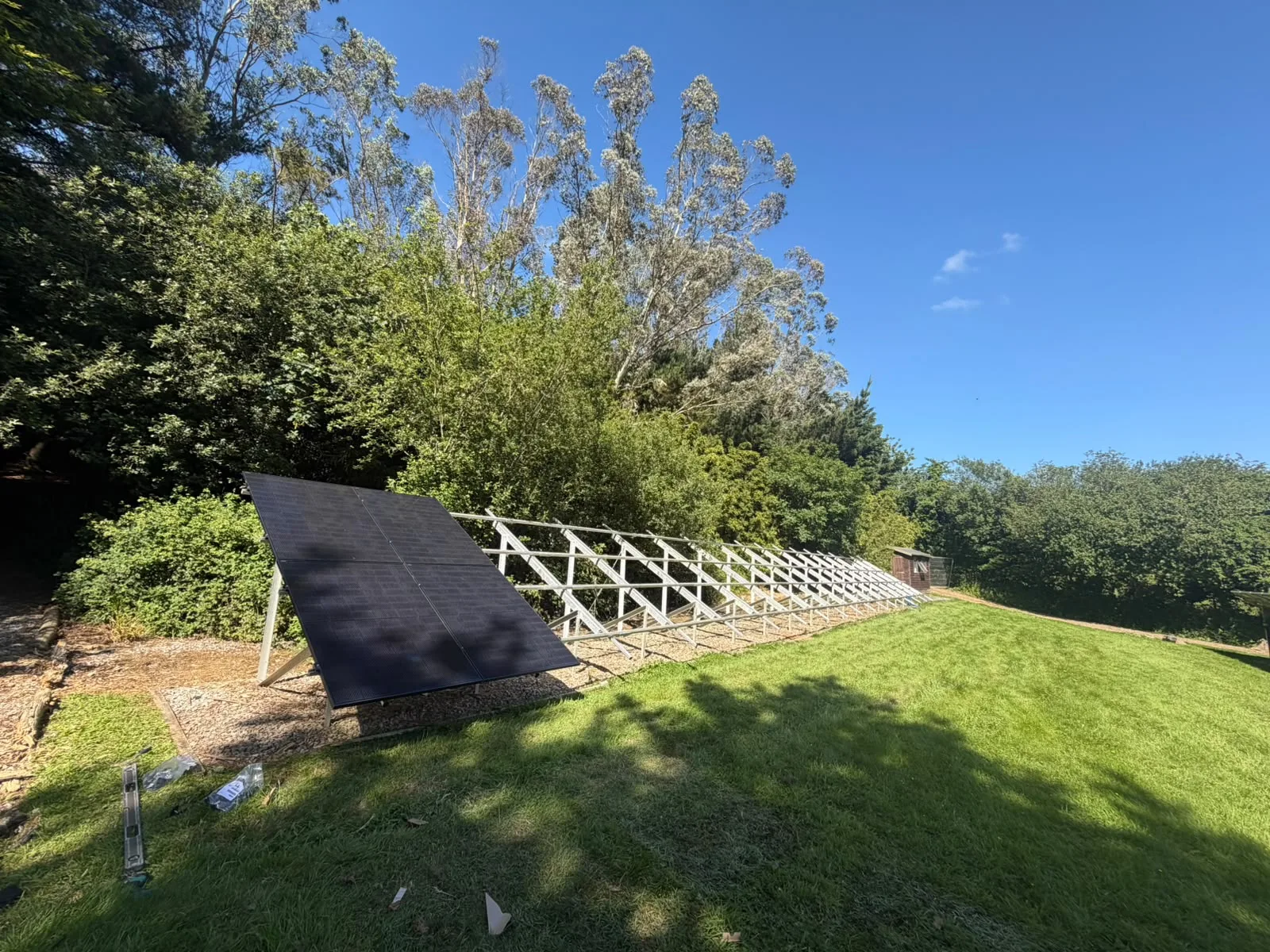 A row of angled metal mounting frames beside ground-mounted solar panels on a lawn, with trees behind under a blue sky.