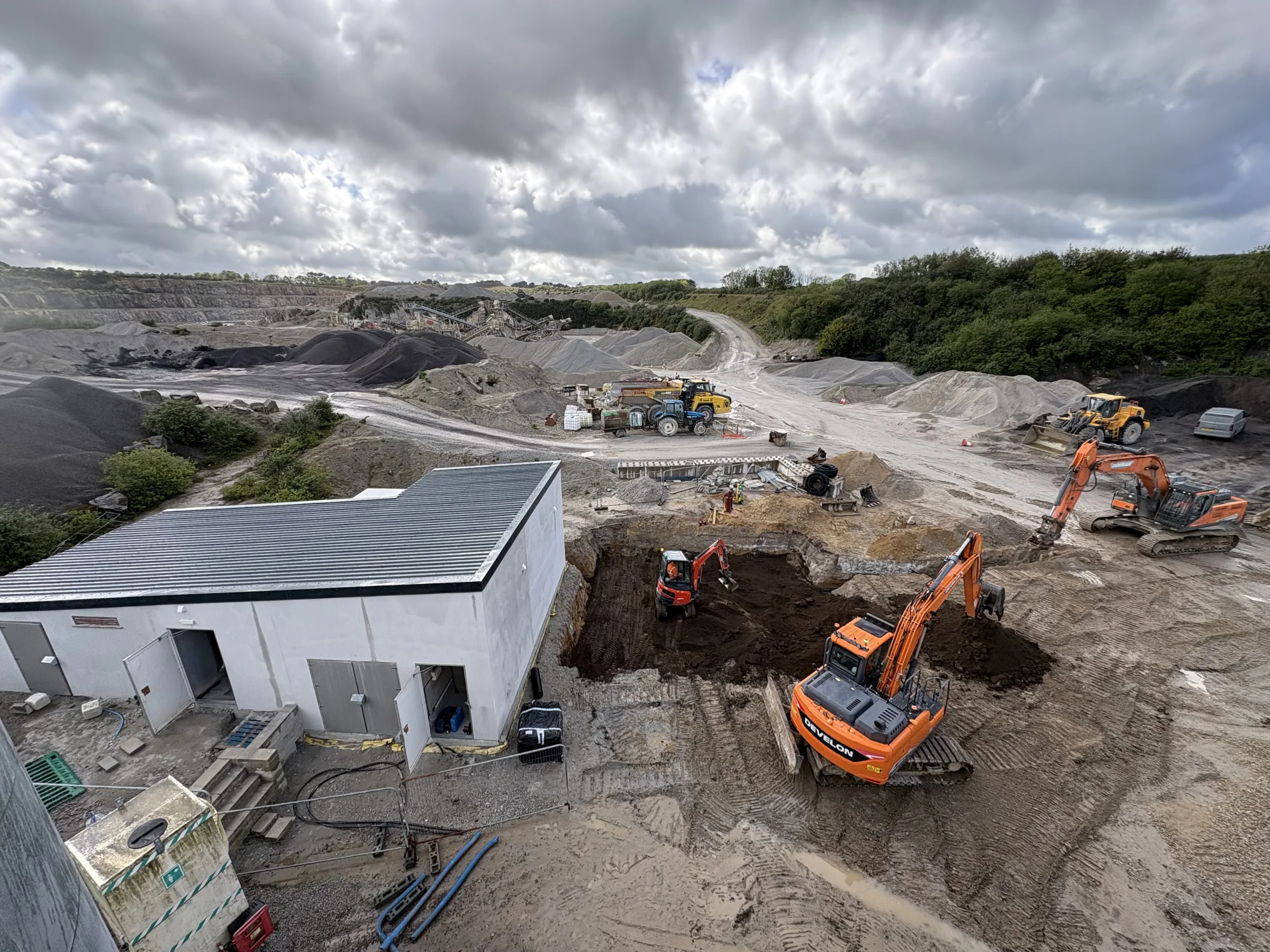 Elevated view of three orange excavators working in dug ground beside a grey industrial building, with aggregate stockpiles and plant beyond.