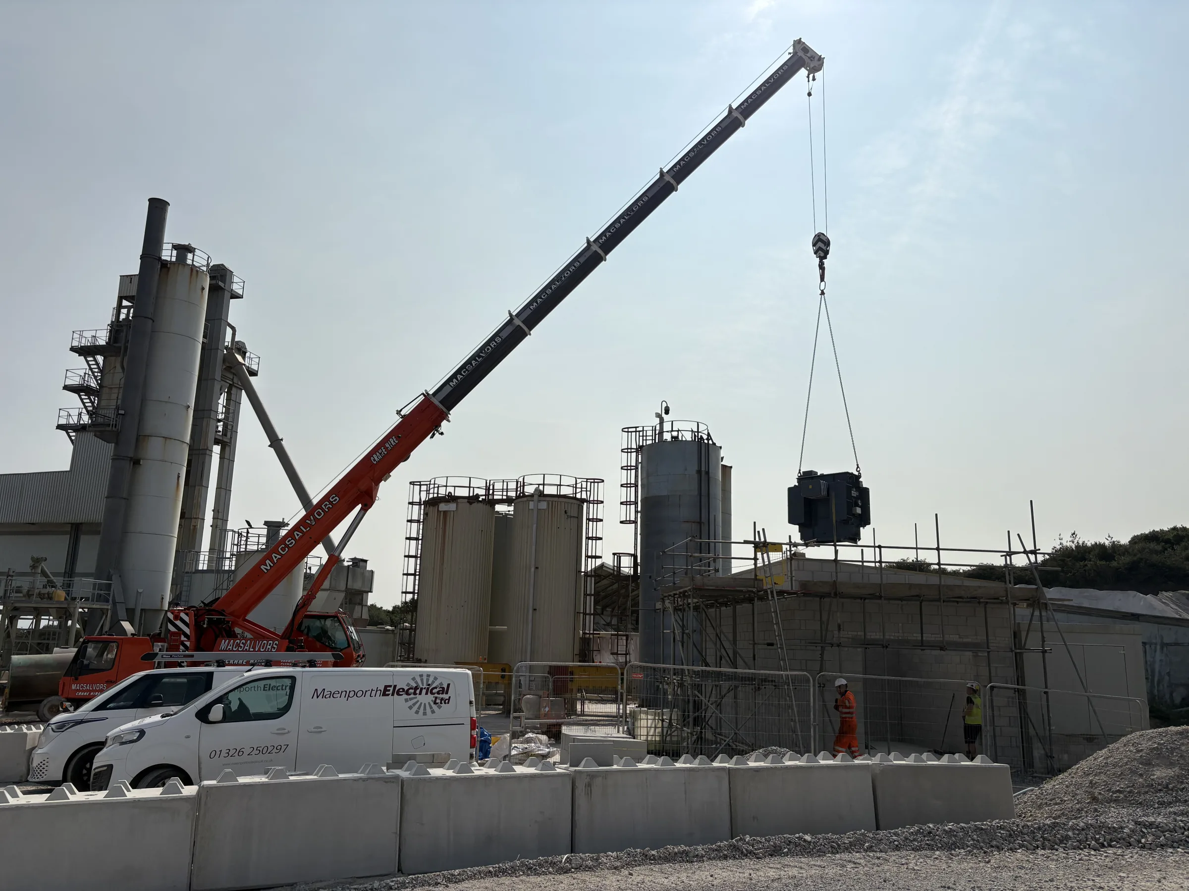 Mobile crane lifting a large grey transformer beside scaffolding and industrial processing structures, with vans and concrete barriers in the foreground.