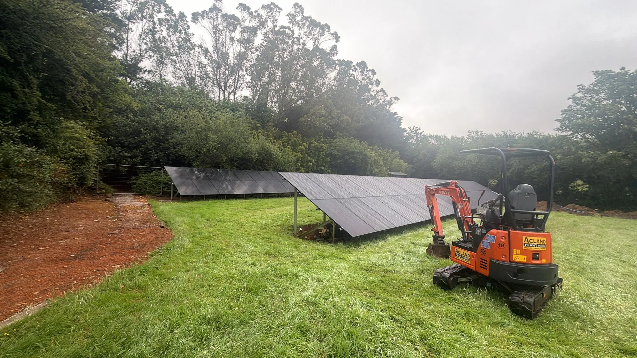 Two rows of dark ground-mounted solar panels on grass, with an orange tracked mini excavator parked beside the nearer row and trees behind.