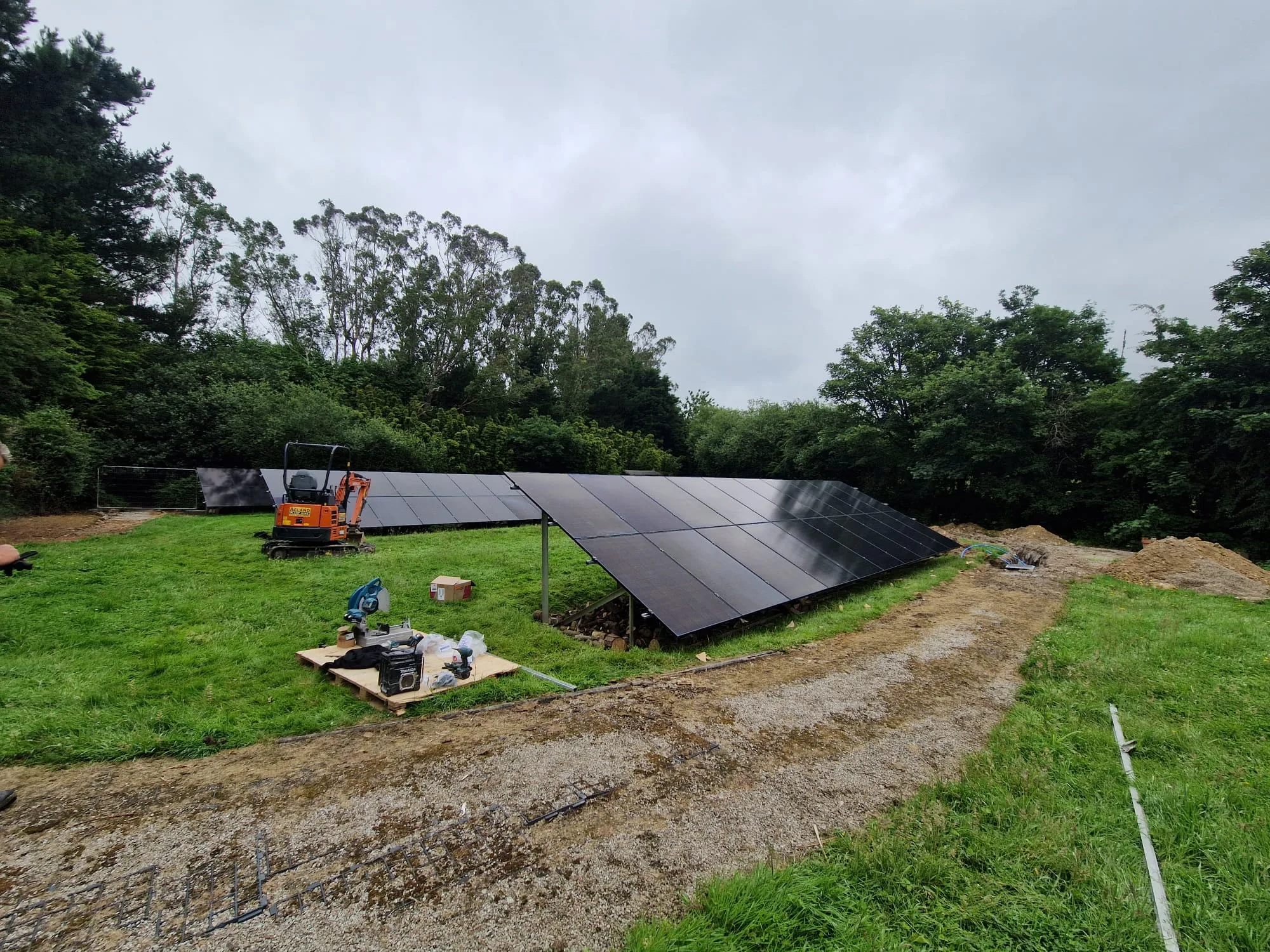 Several rows of ground-mounted solar panels in a grassy clearing, with a small orange excavator, tools and a curving gravel track.