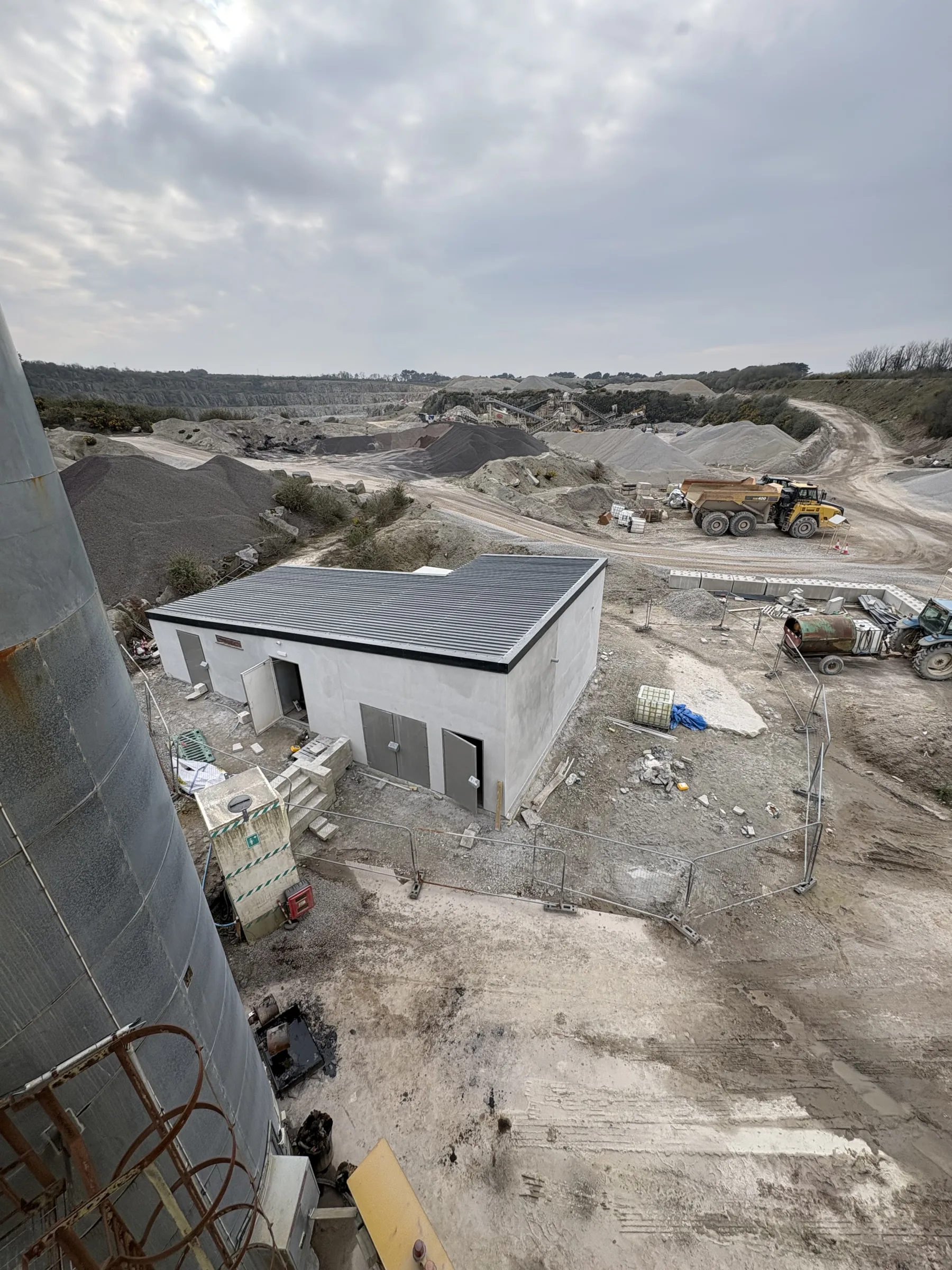 Elevated view over a quarry site with a small white building, aggregate piles, haul vehicles and temporary fencing.
