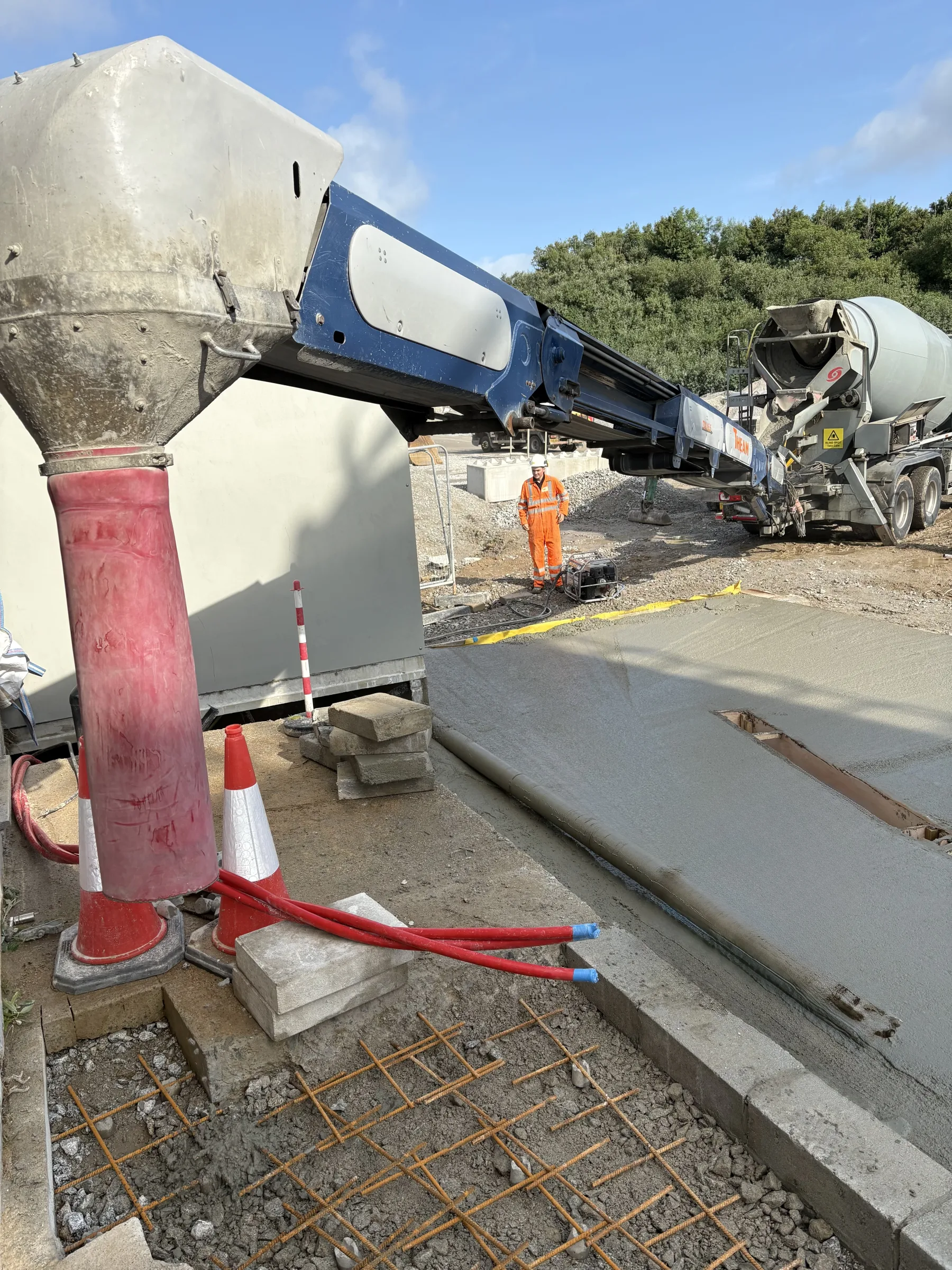 Concrete pump boom positioned beside a mixer lorry, with a worker in orange high-visibility clothing near a freshly poured slab.