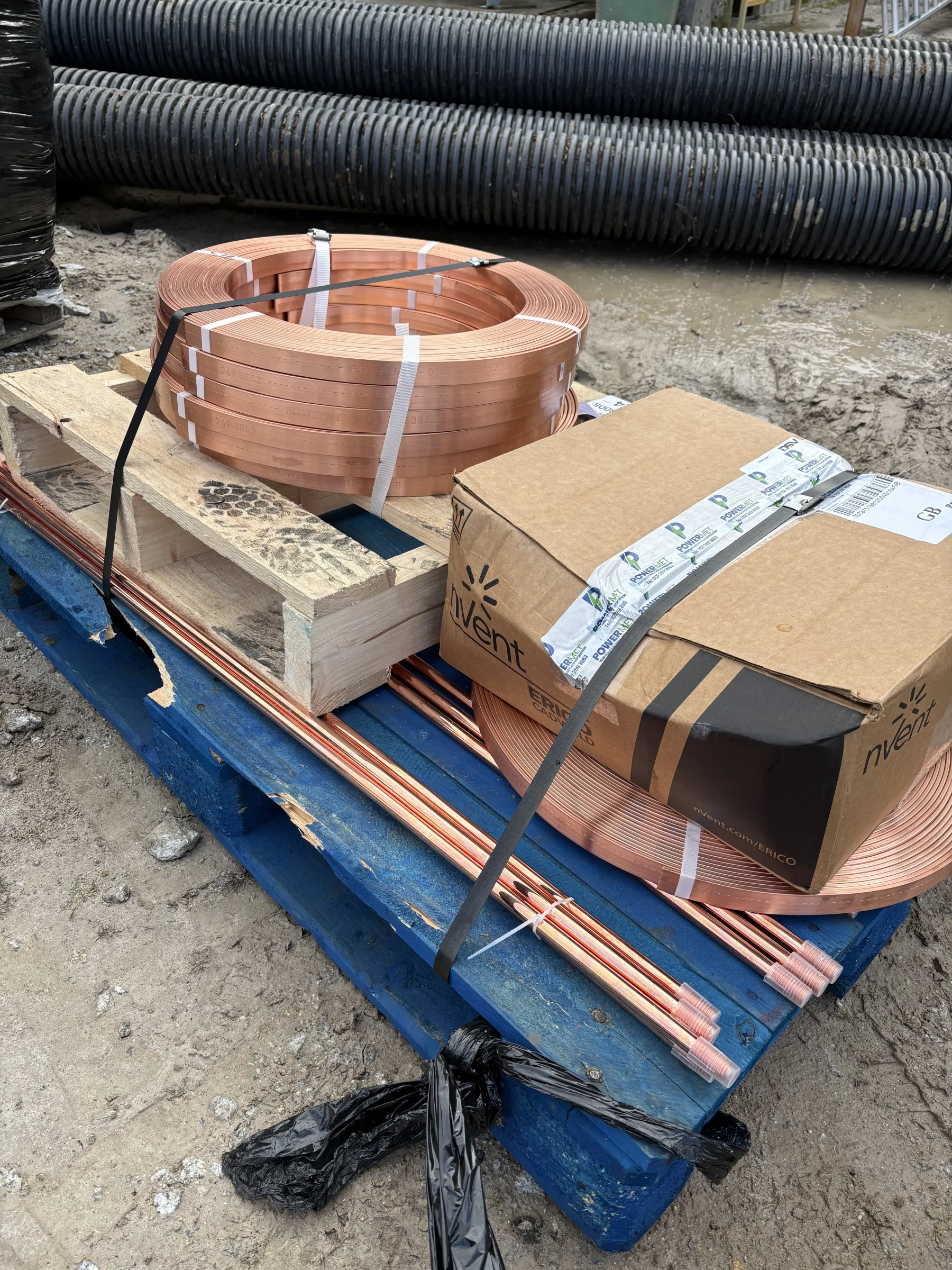 Coils of perforated copper tape, straight copper lengths, timber boards and a cardboard box strapped to a blue pallet, with black corrugated pipes behind.