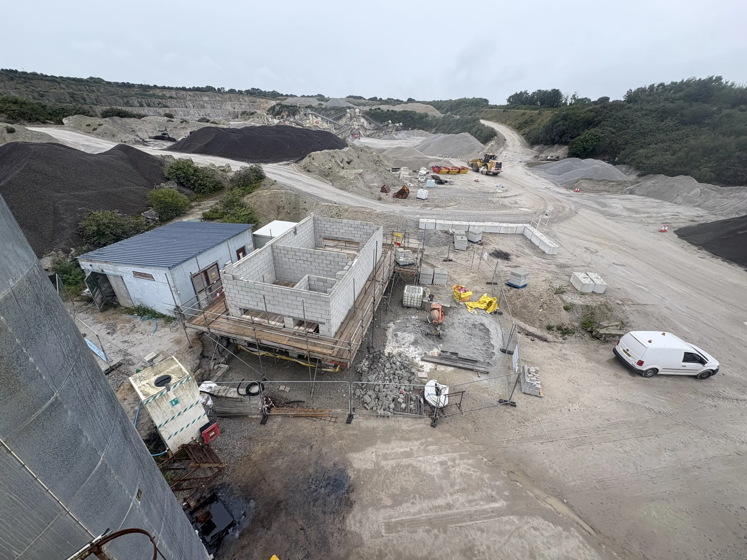 High-angle view across a quarry construction area with an unfinished block building, temporary fencing, aggregate piles, machinery and a white van.