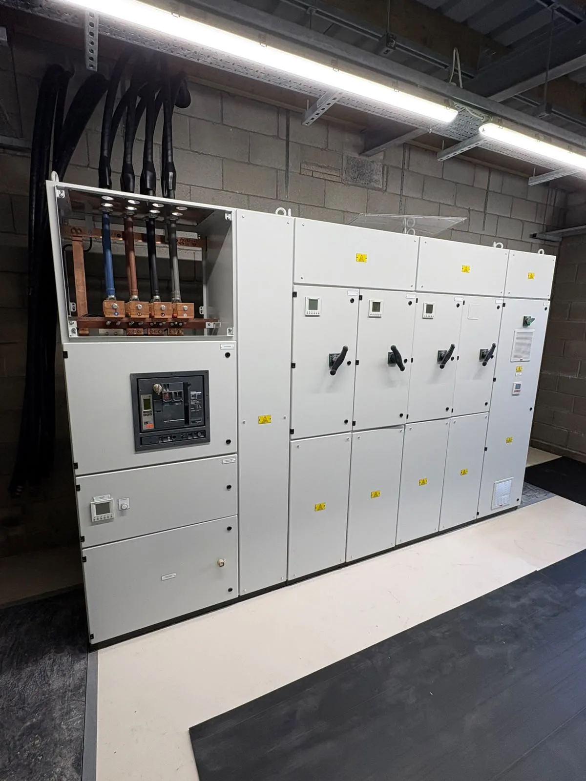 Large grey electrical switchgear cabinet with multiple compartment doors, black operating handles, an exposed upper busbar section and heavy cables entering from above.