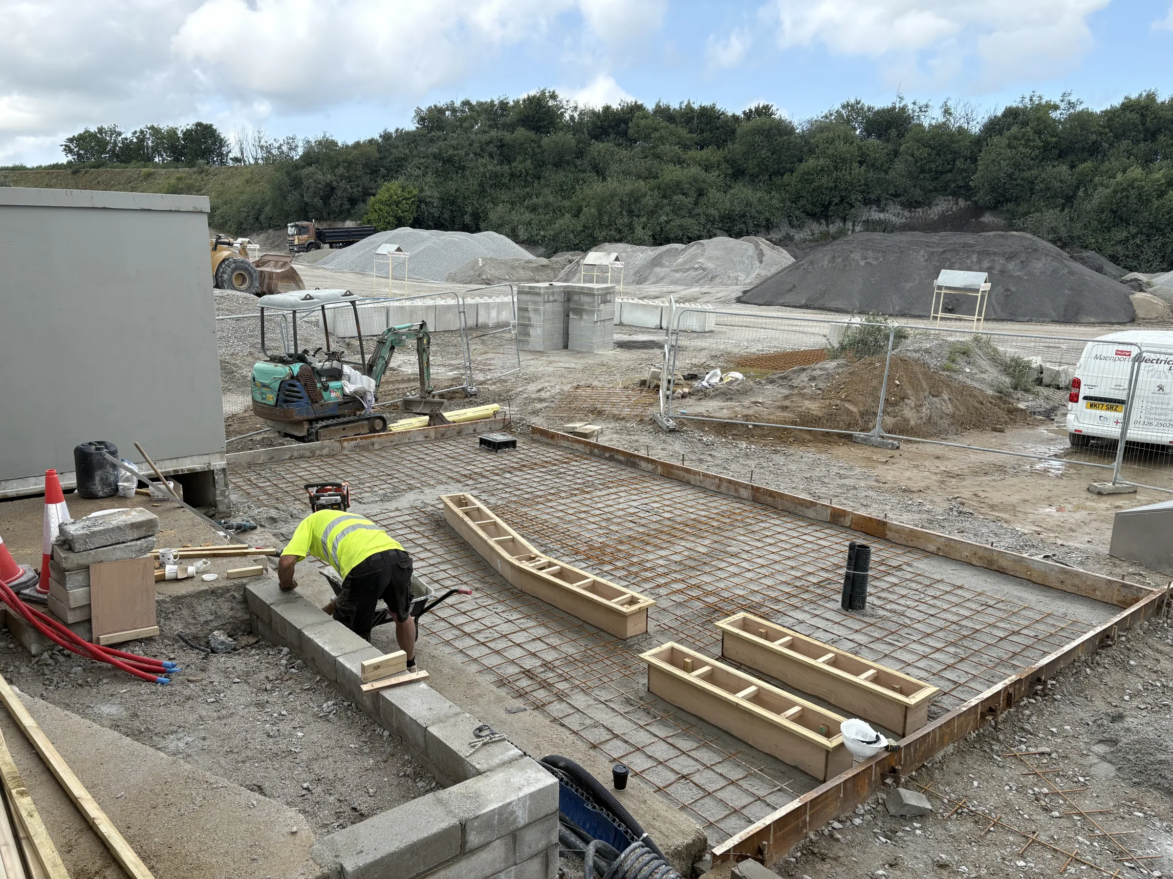 Worker in yellow high-visibility clothing kneeling beside a block edge around a reinforced foundation slab with timber formwork.