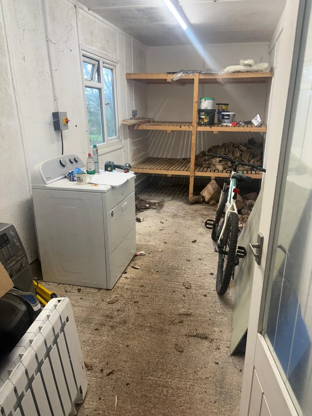 Utility room containing a washing machine, portable radiator, bicycle and timber shelving loaded with logs, with a bright ceiling strip light overhead.