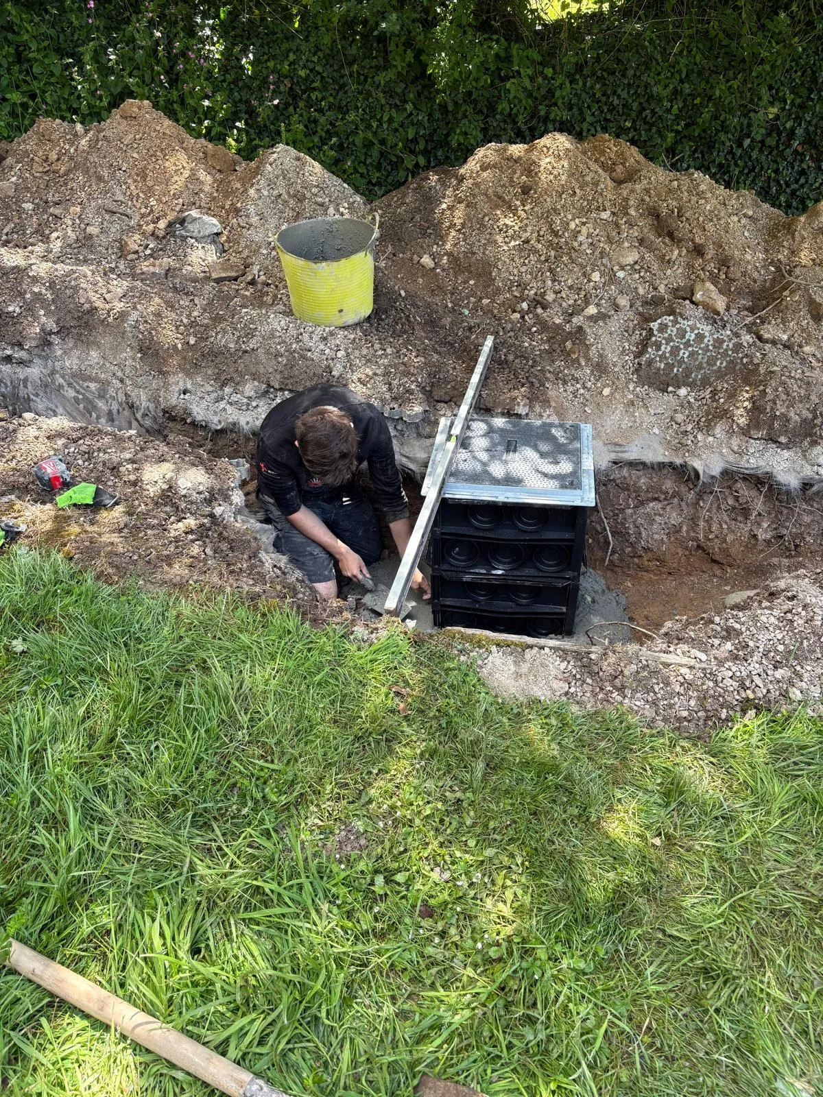 Worker kneeling in an excavated trench beside a black underground chamber, with a spirit level, bucket and spoil piles nearby.