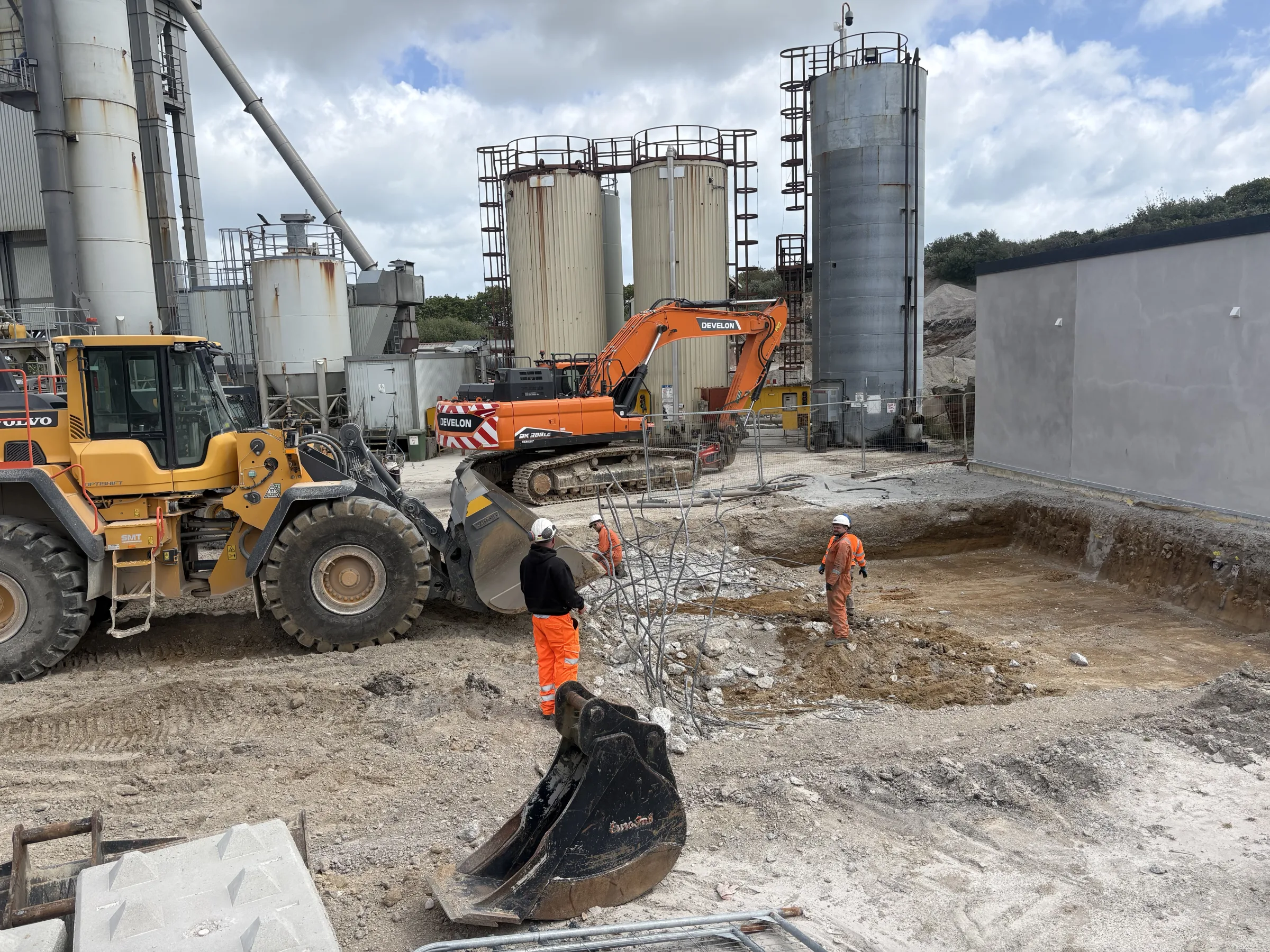 Construction area with a yellow wheel loader, orange tracked excavator, two workers in high-visibility clothing and tall industrial tanks beside a grey building.