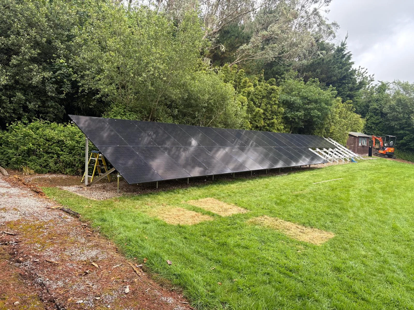 Long row of dark solar panels on a low ground-mounted frame beside a grass lawn and dense trees.