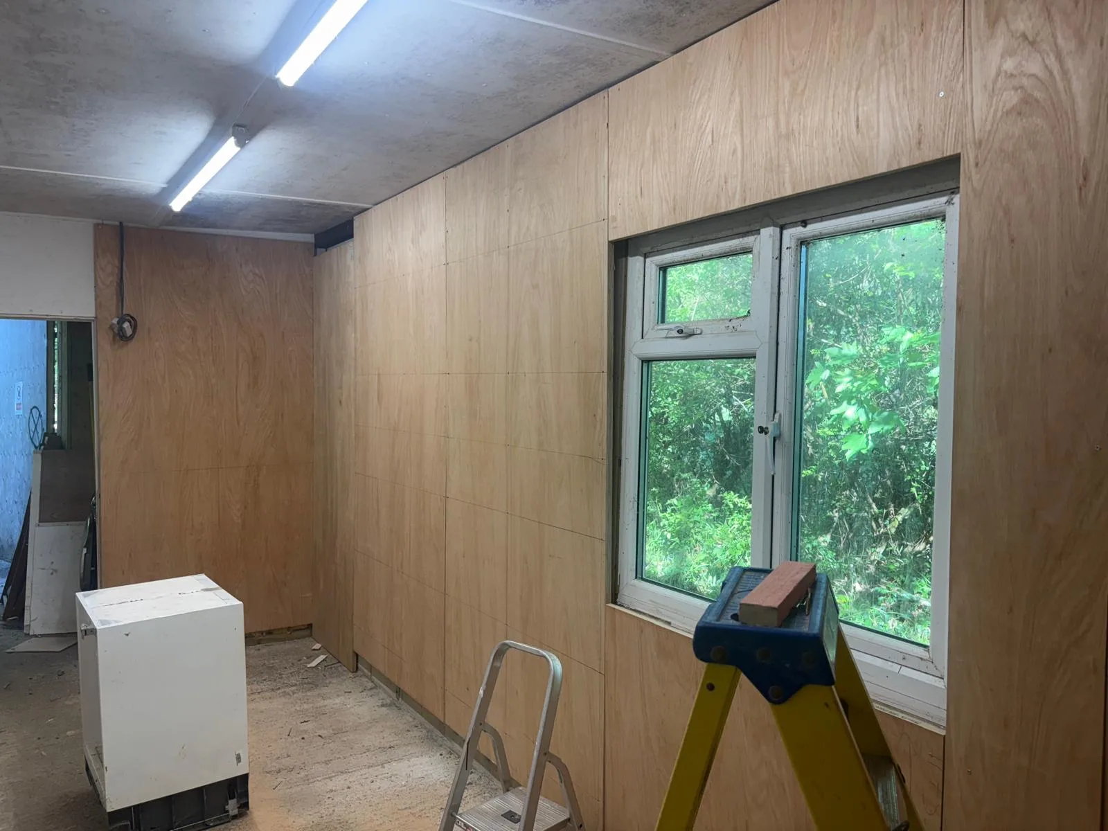 Interior room with plywood-lined walls, two illuminated ceiling battens, a large window, a yellow step ladder and a white cabinet.