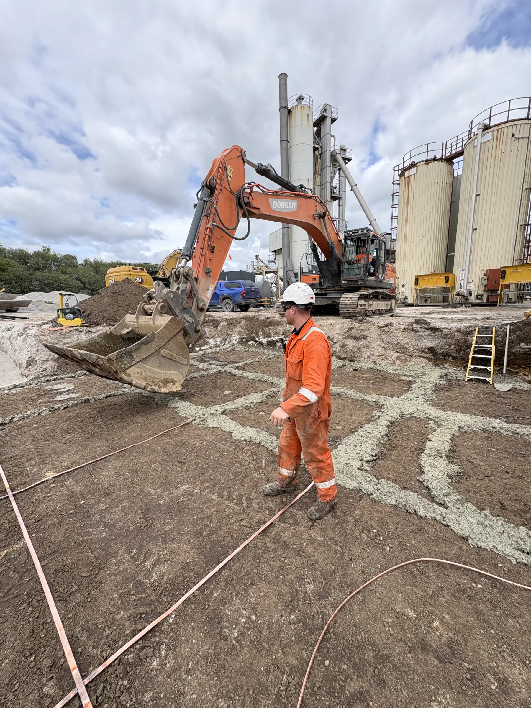 Worker in orange protective clothing and a white hard hat standing among marked lines on prepared ground, with a tracked excavator and industrial tanks behind.