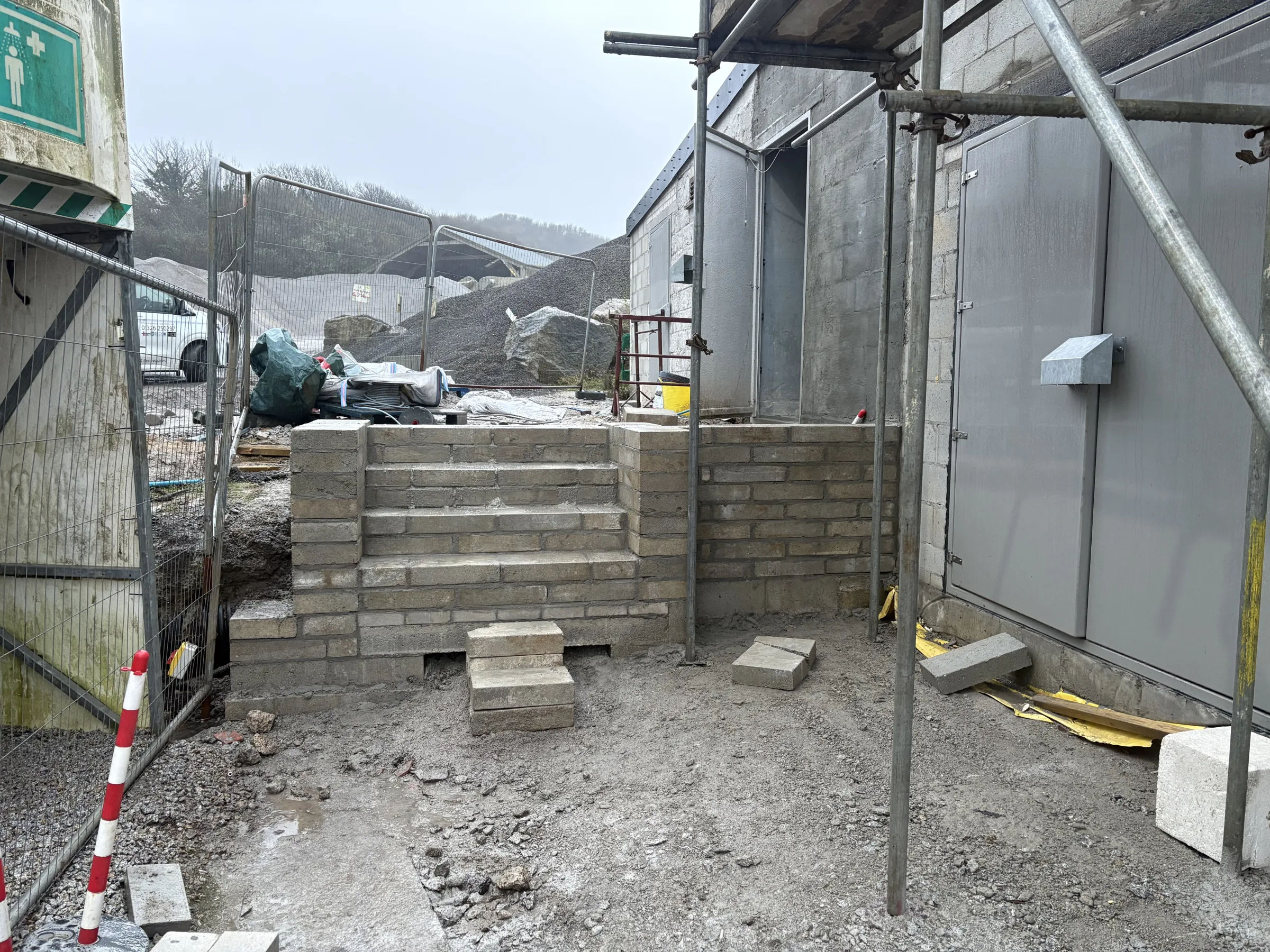 Gravelled work area beside industrial buildings with scaffolding poles, temporary fencing, a low block wall and a grey external enclosure.