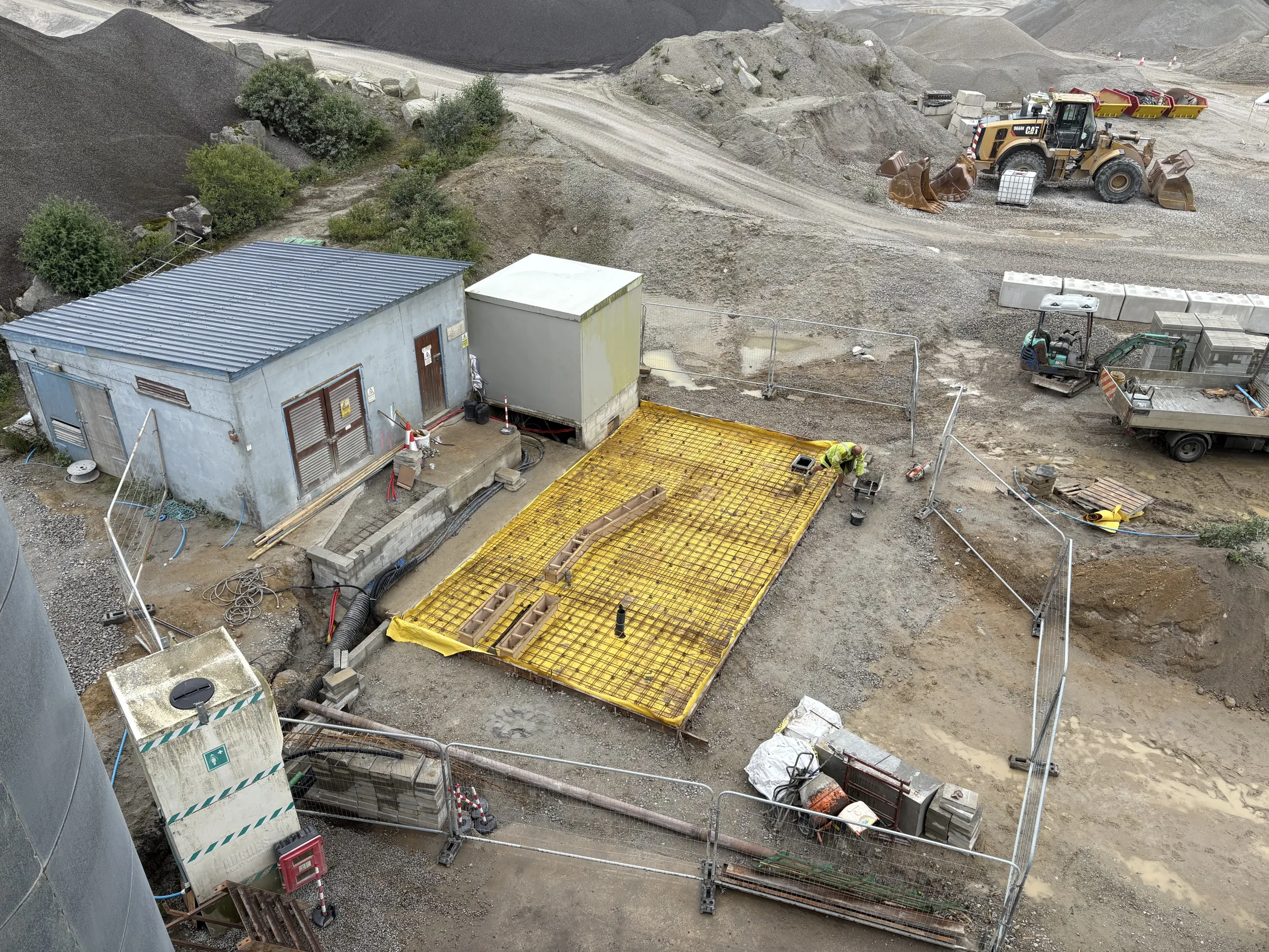 Elevated view of a rectangular yellow-covered slab with steel reinforcement beside a small building and site enclosure.