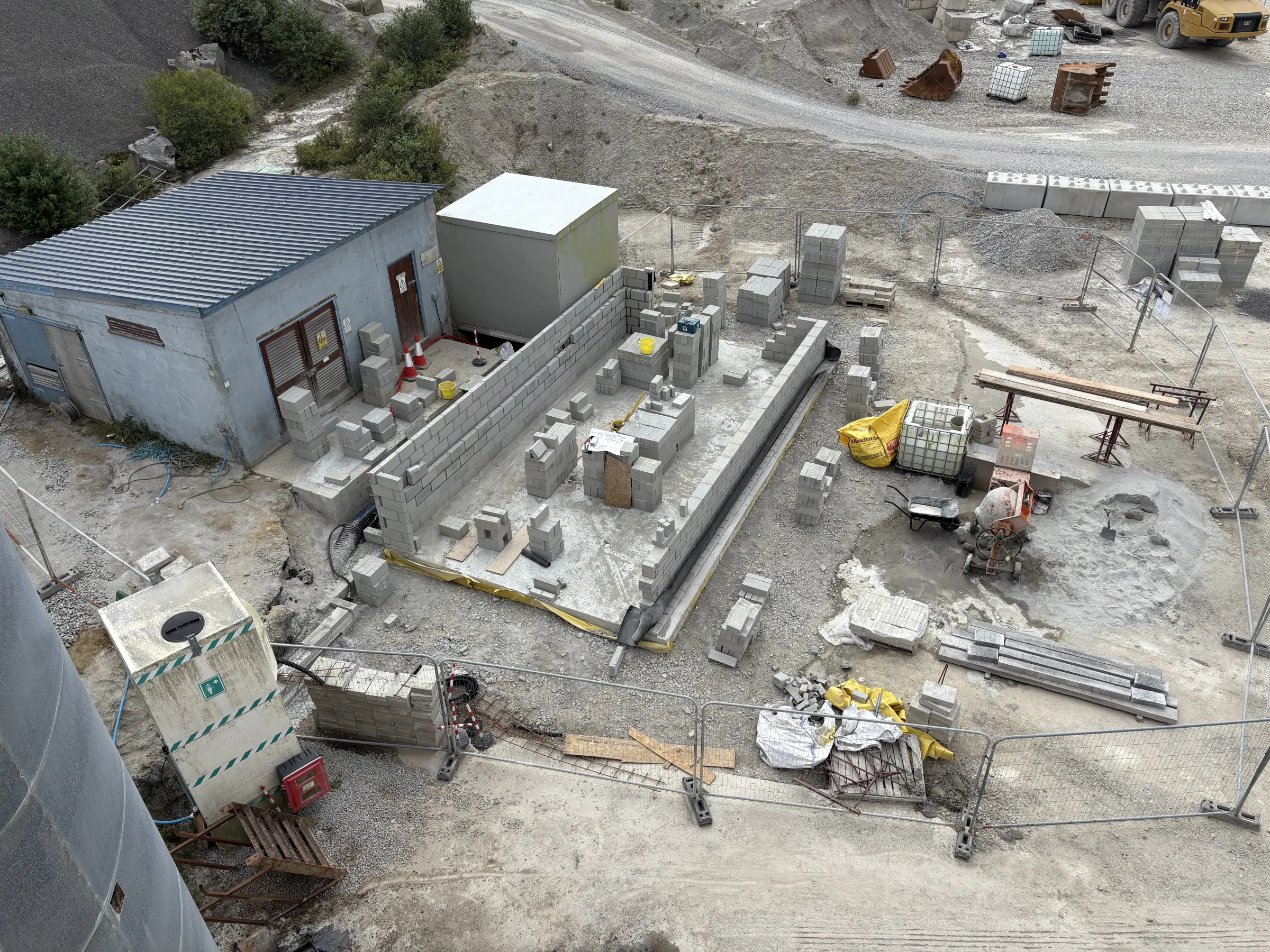 High-angle view of a rectangular blockwork structure under construction beside a small building, with blocks, tools, fencing and materials spread around the site.