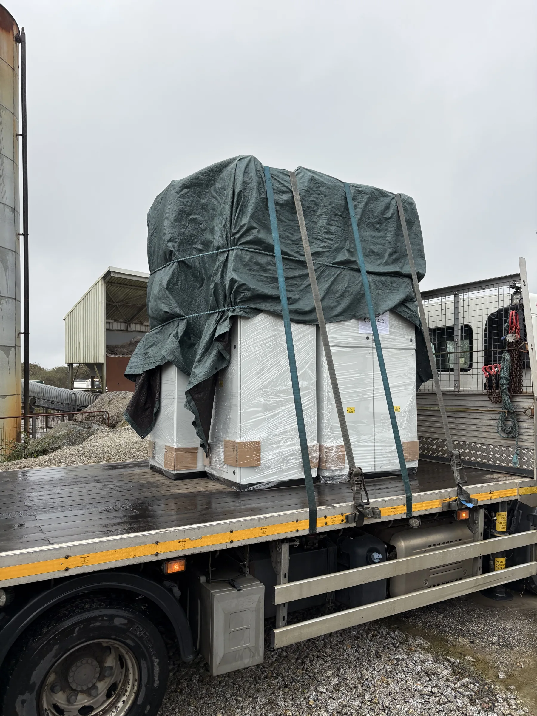 Large wrapped equipment secured with blue straps on the rear of a flatbed lorry in an industrial yard.