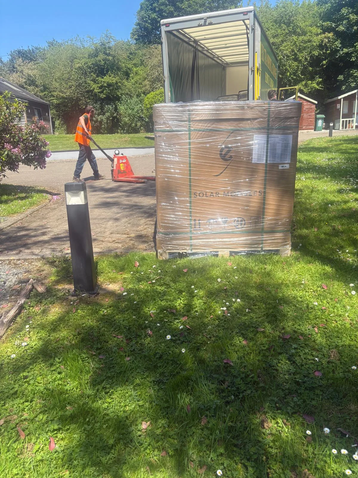Large shrink-wrapped pallet on grass behind an open-backed lorry, with a worker in orange high-visibility clothing moving a pallet truck nearby.