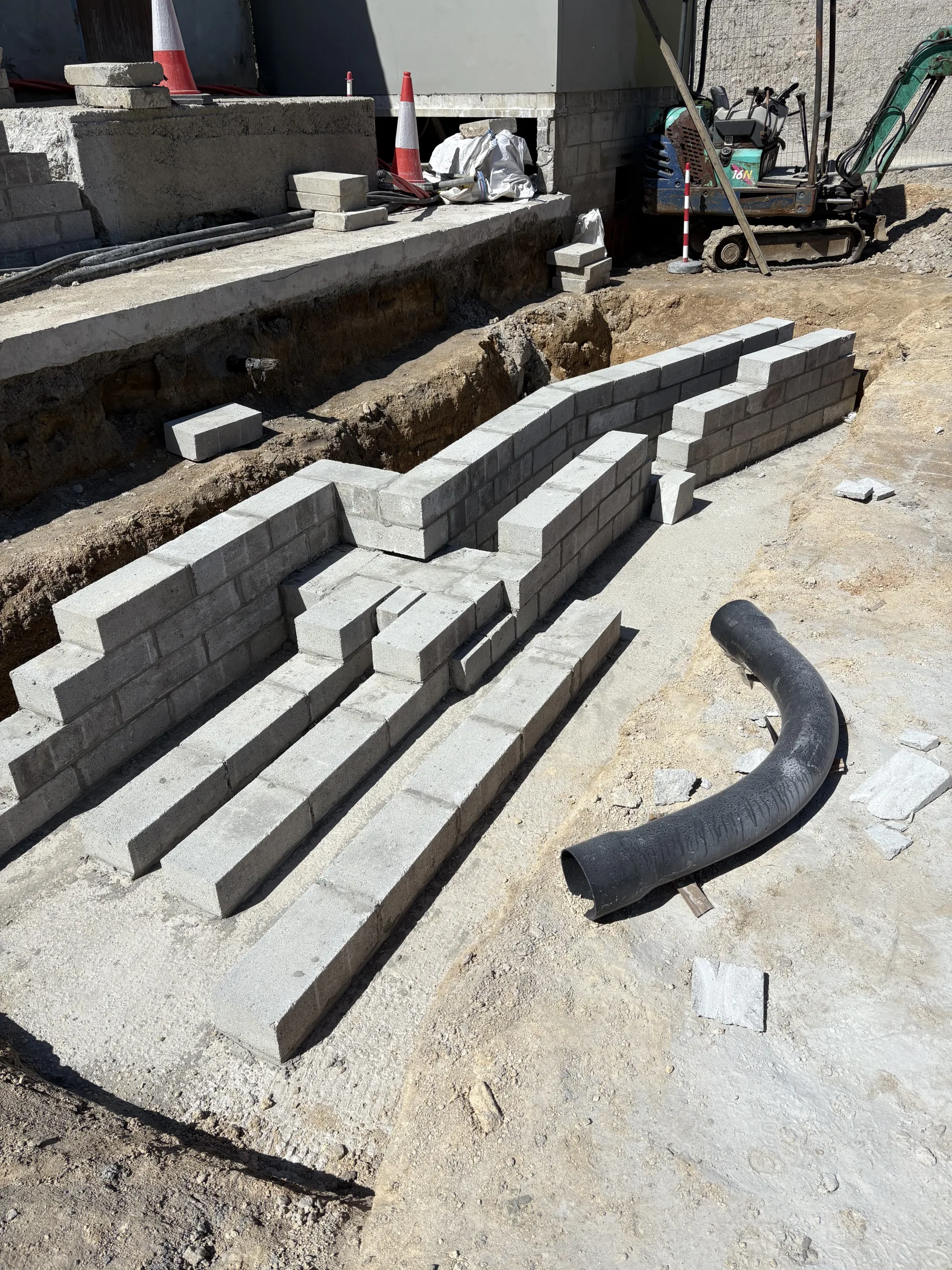 Rows of grey concrete blocks forming narrow channels beside an open trench, with timber lengths and a curved black pipe in the foreground.