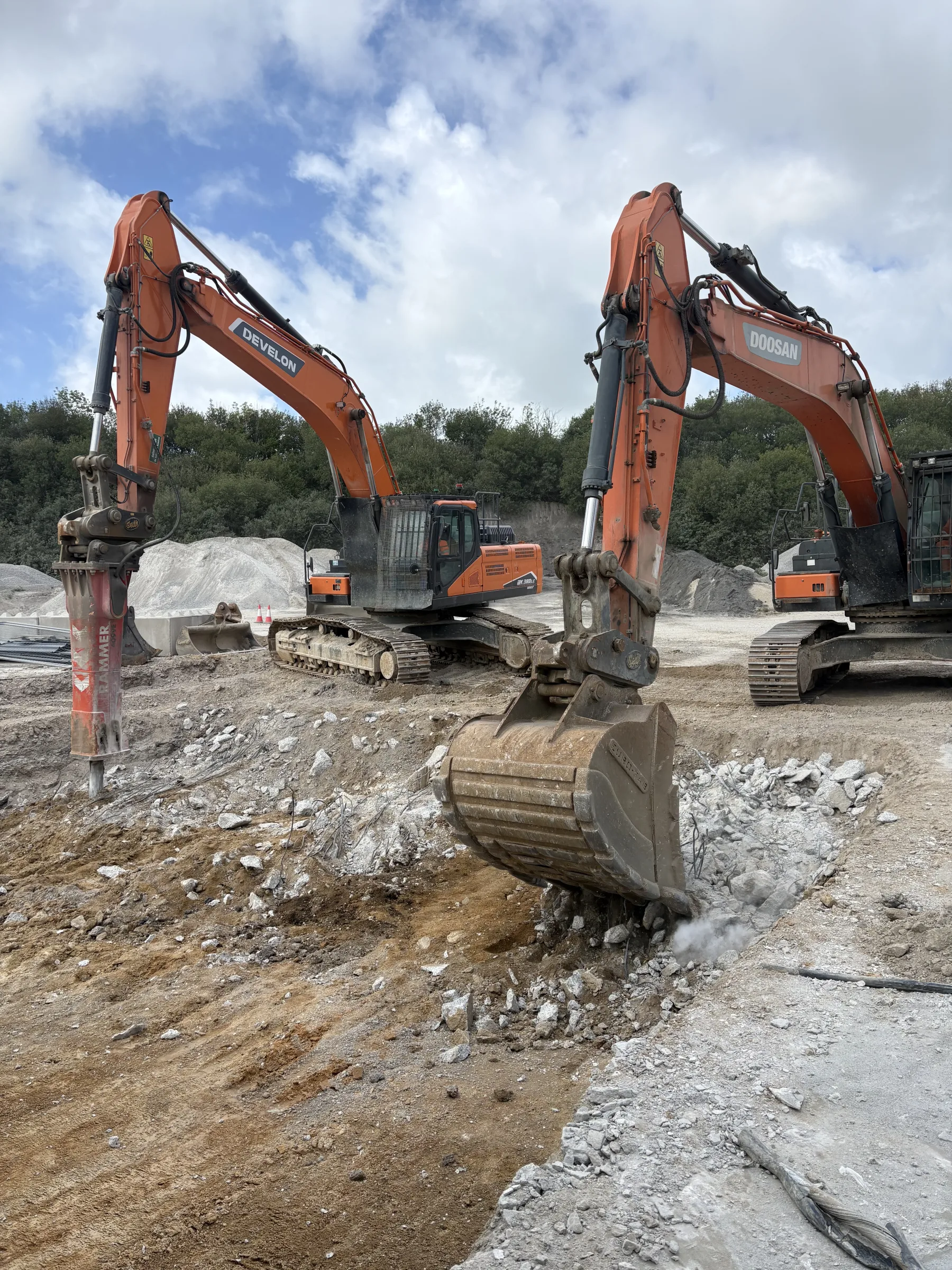 Two orange tracked excavators stand in rocky ground, one fitted with a hydraulic breaker and the other with a digging bucket.