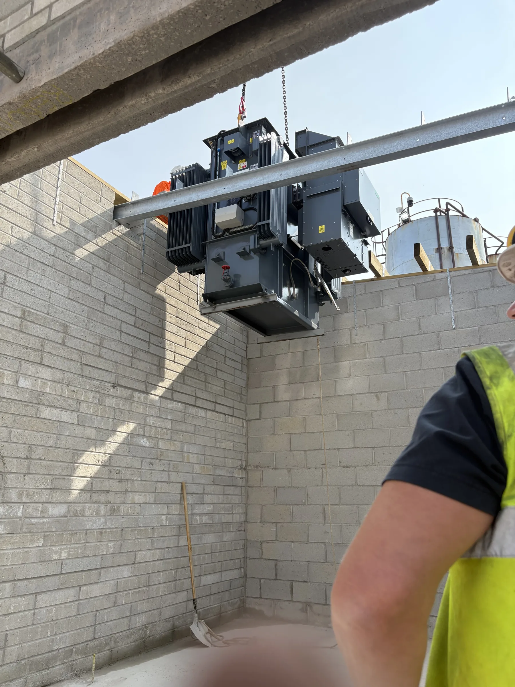 Large grey electrical transformer suspended from chains above an unfinished concrete block enclosure, with part of a worker in a high-visibility vest at right.